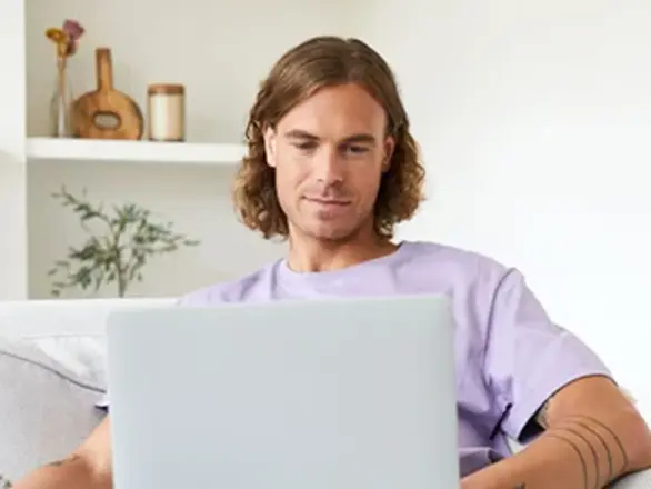 A person with long hair and a tattoo sits on a sofa, looking at a laptop in a bright, modern living room.