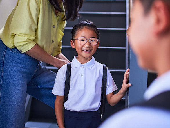 A child wearing glasses and a school uniform stands on stairs, smiling, with an adult behind and another child in the foreground.