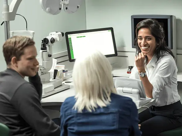 An eye care professional smiles while talking to two patients in an examination room with medical equipment and computer monitors.