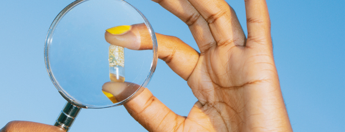 Image of a hand holding up a magnifying glass to an Essential for Women 18+ capsule with a blue background.