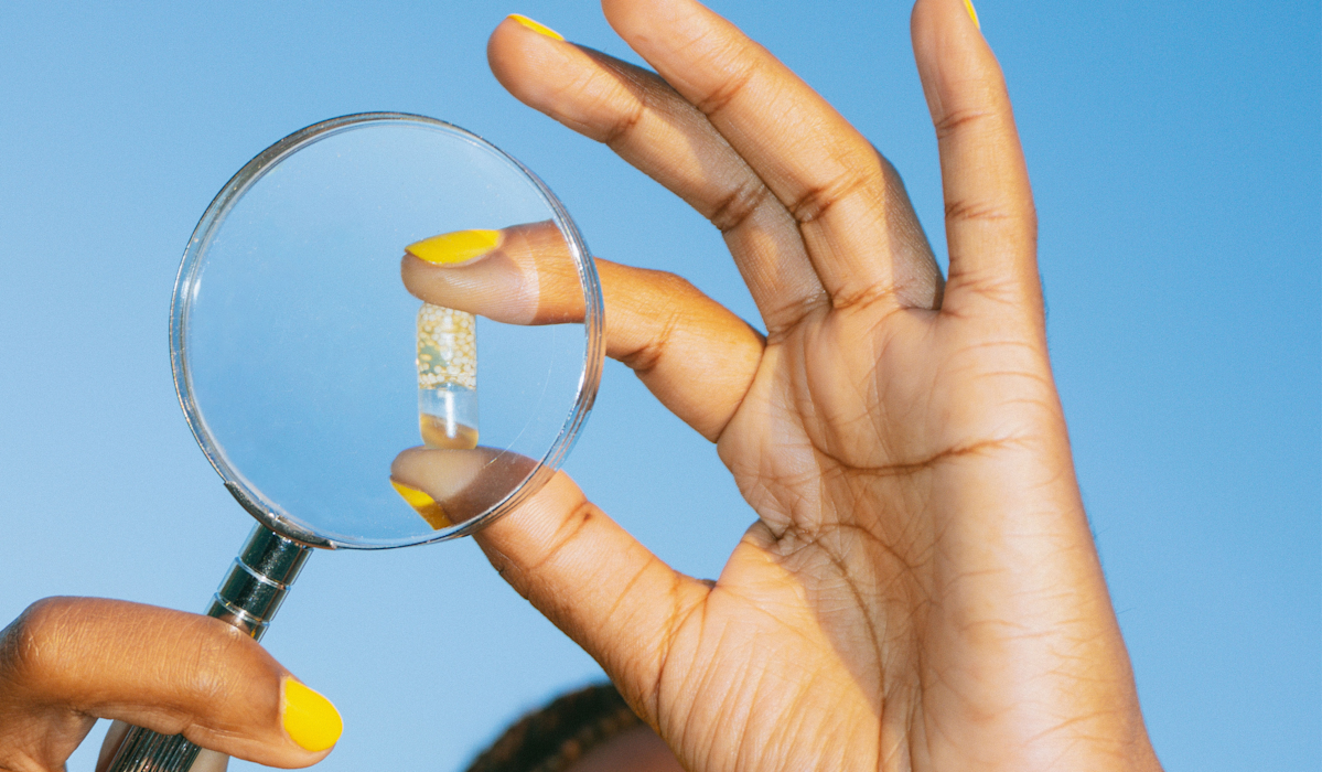 Image of a hand holding up a magnifying glass to an Essential for Women 18+ capsule with a blue background.