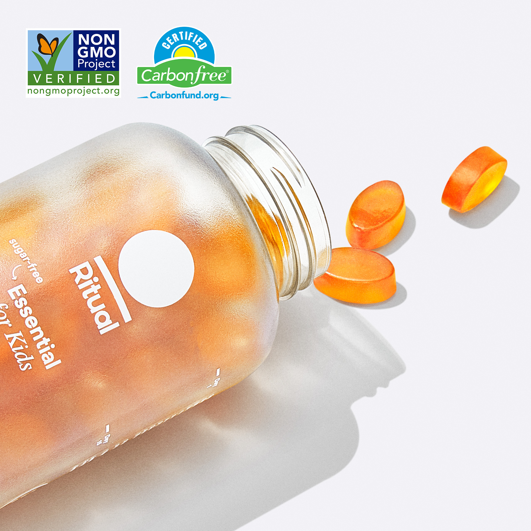 A close up image of the frosted Essential for Kids 4+ bottle. It is on its side with the cap off and three (3) Essential for Kids 4+ multivitamin gummies are spilling out. The gummies are oval and orange in color.