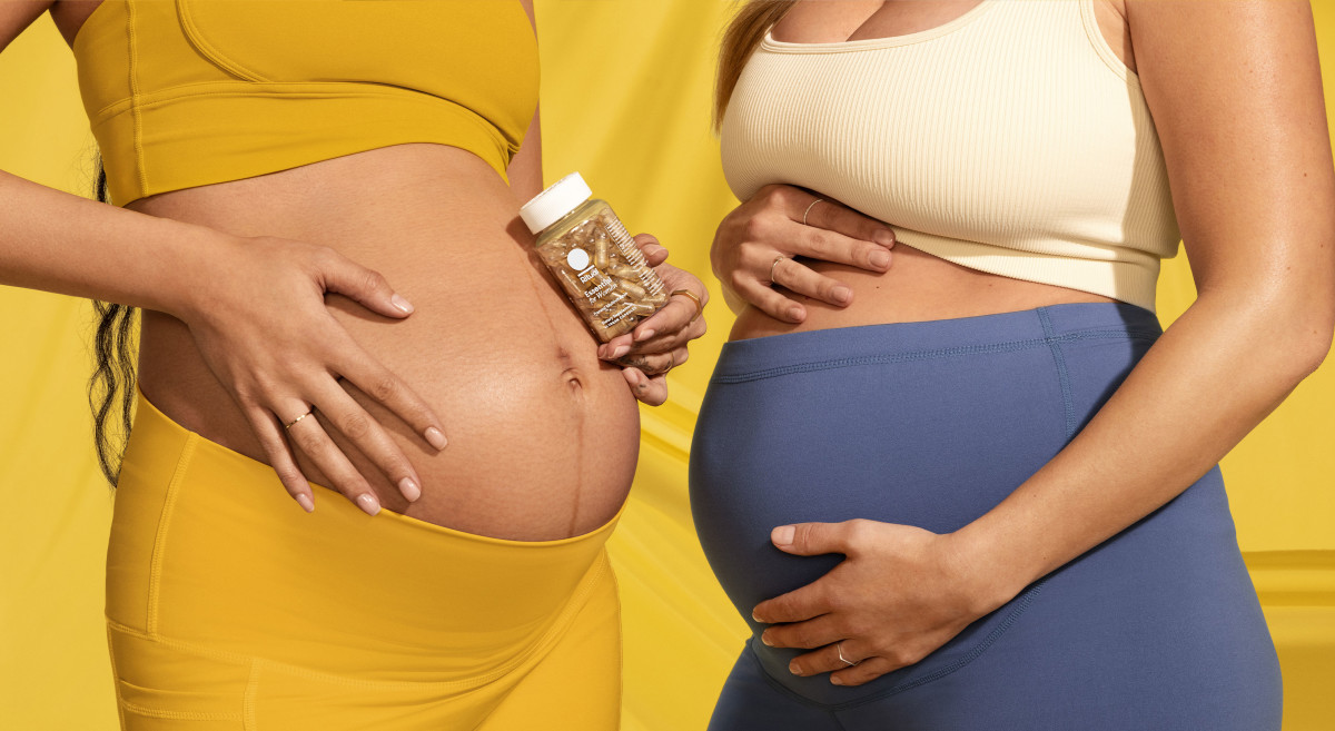 Two pregnant models standing next to each other with one holding a bottle of Essential Prenatal with a yellow background.