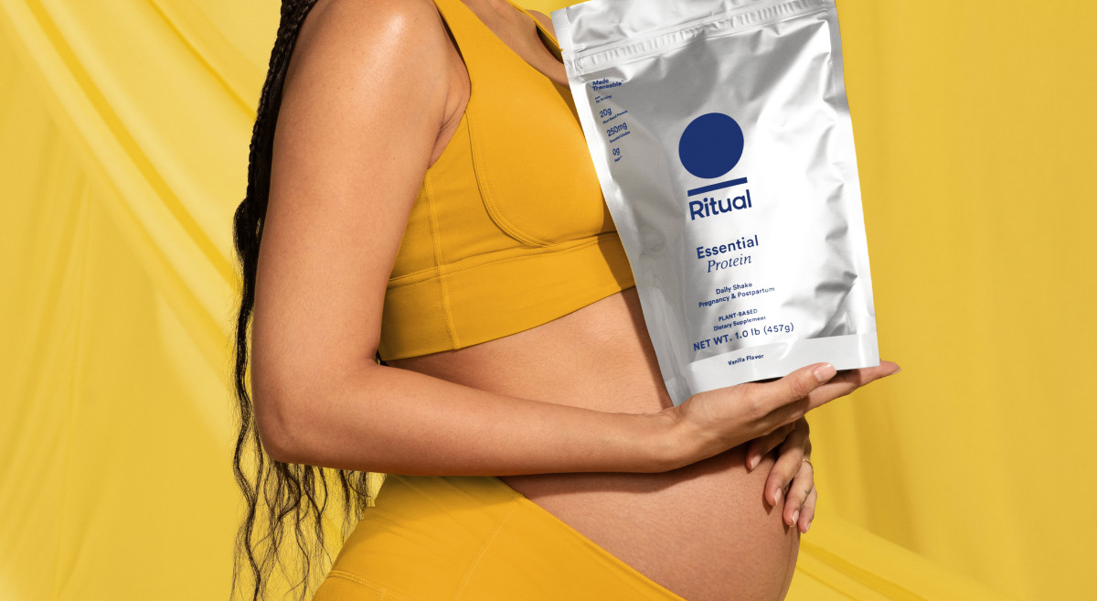 Pregnant model holding Essential Protein Daily Shake Pregnancy & Postpartum package with yellow background.