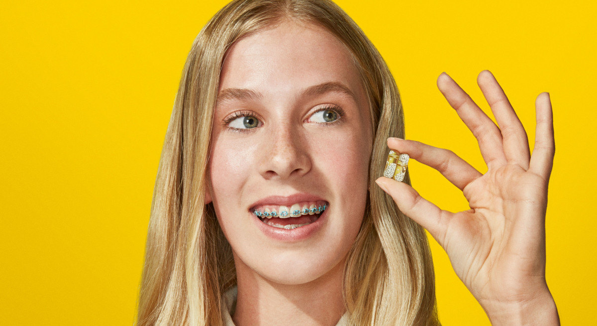 Teenage model smiling and holding two Ritual multivitamin capsules in her hand.