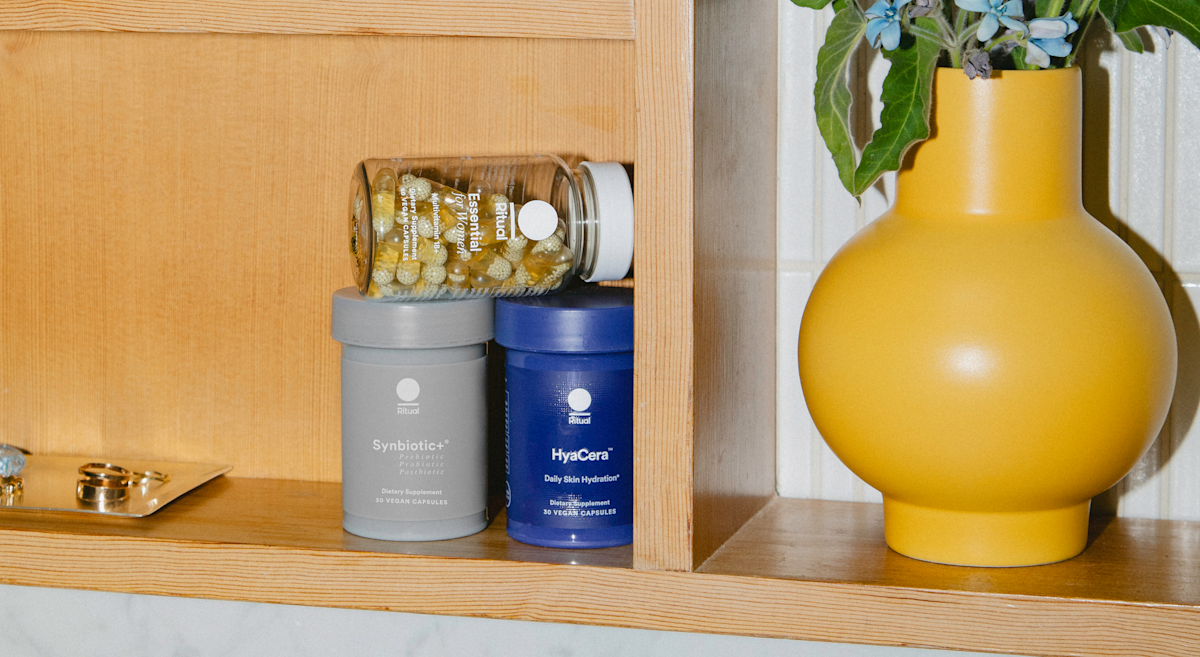 A bottle of Essential for Women 18+, Synbiotic+ and Hyacera on a shelf next to a yellow vase.