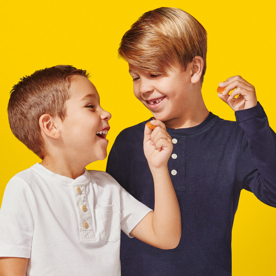 Two young boys smiling and each boy is holding an Essential for Kids 4+ multivitamin gummy.