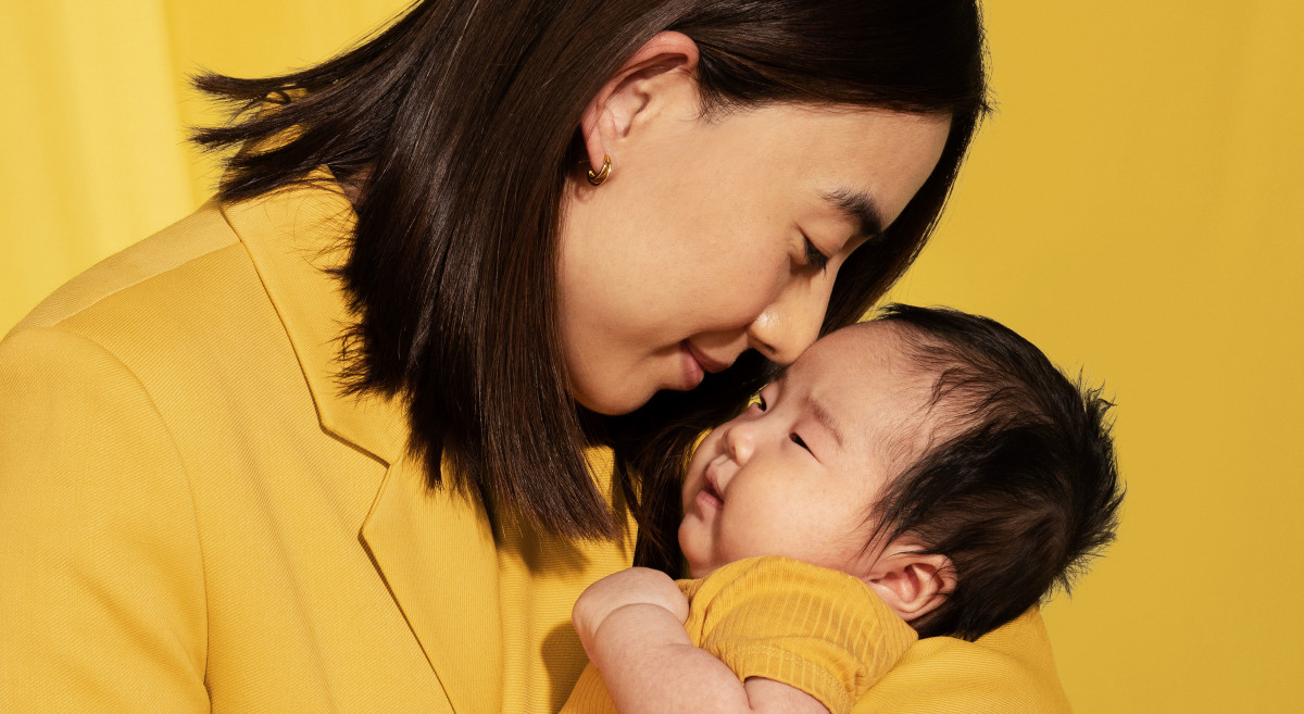 A model holding a baby and touching her nose to the baby's forehead with a yellow background.