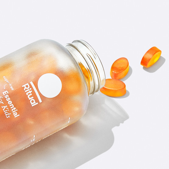 A close up image of the frosted Essential for Kids 4+ bottle. It is on its side with the cap off and three (3) Essential for Kids 4+ multivitamin gummies are spilling out. The gummies are oval and orange in color.