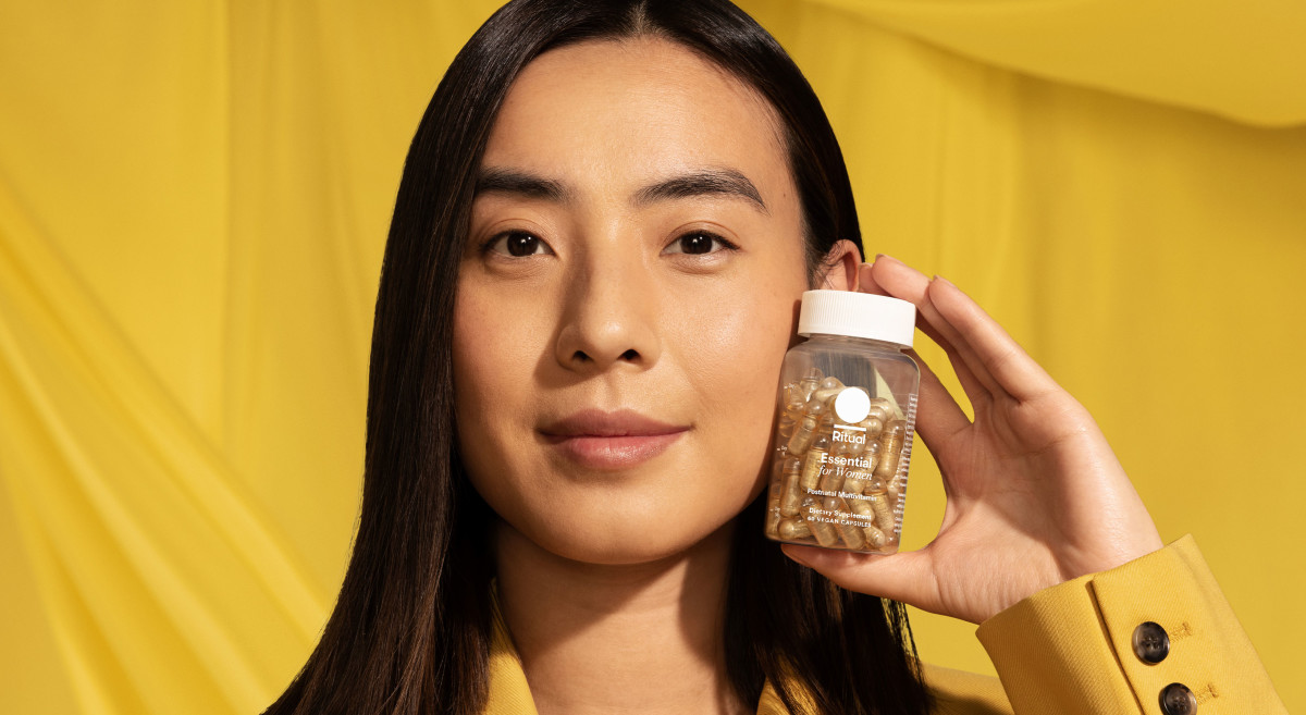 Model holding a bottle of Essential Postnatal Multivitamin next to her face with a yellow background.
