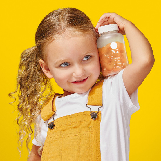 A little girl with blonde hair smiling and holding the Essential for Women 4+ Multivitamin bottle on her shoulder. The background is yellow and she is wearing yellow overalls with a white shirt underneath.