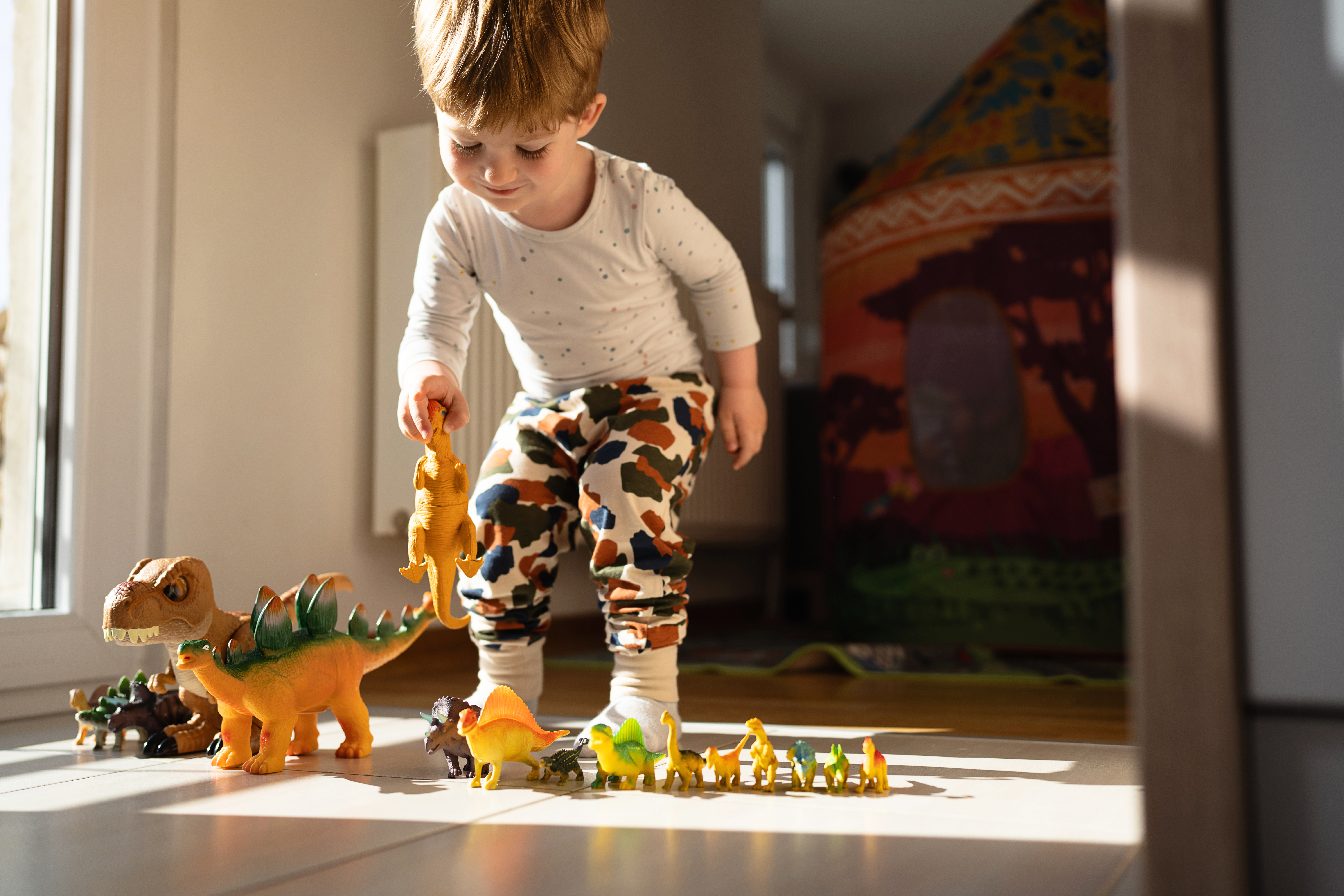 A young child in a white shirt and colorful pants plays with toy dinosaurs on the floor, with sunlight streaming in and a colorful tent in the background.