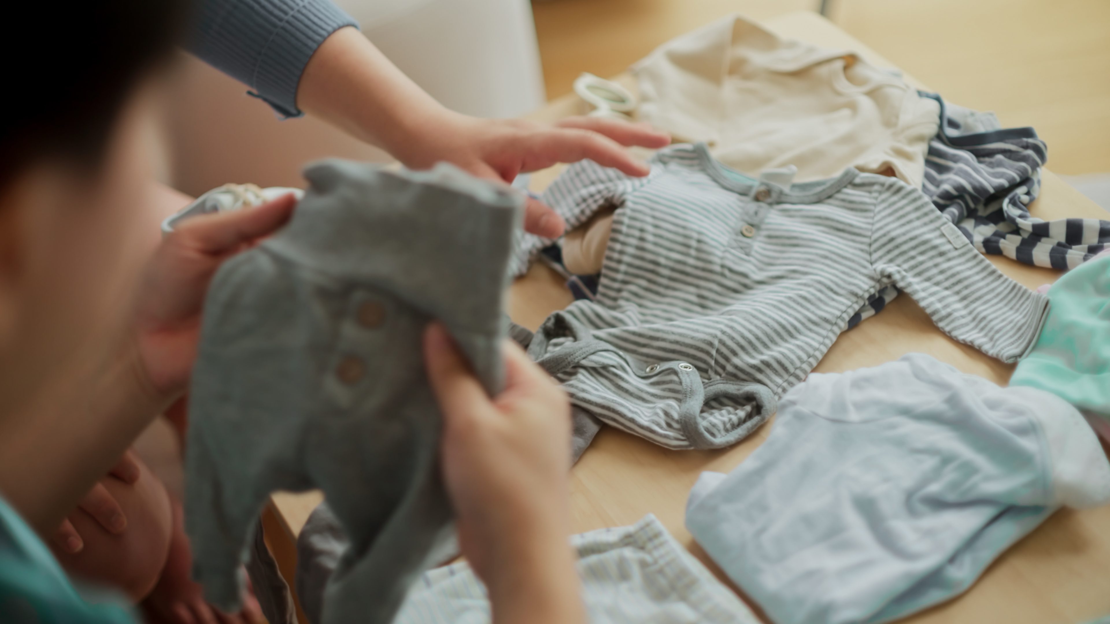 Two people sorting baby clothes on a table, holding a grey onesie and arranging other garments.