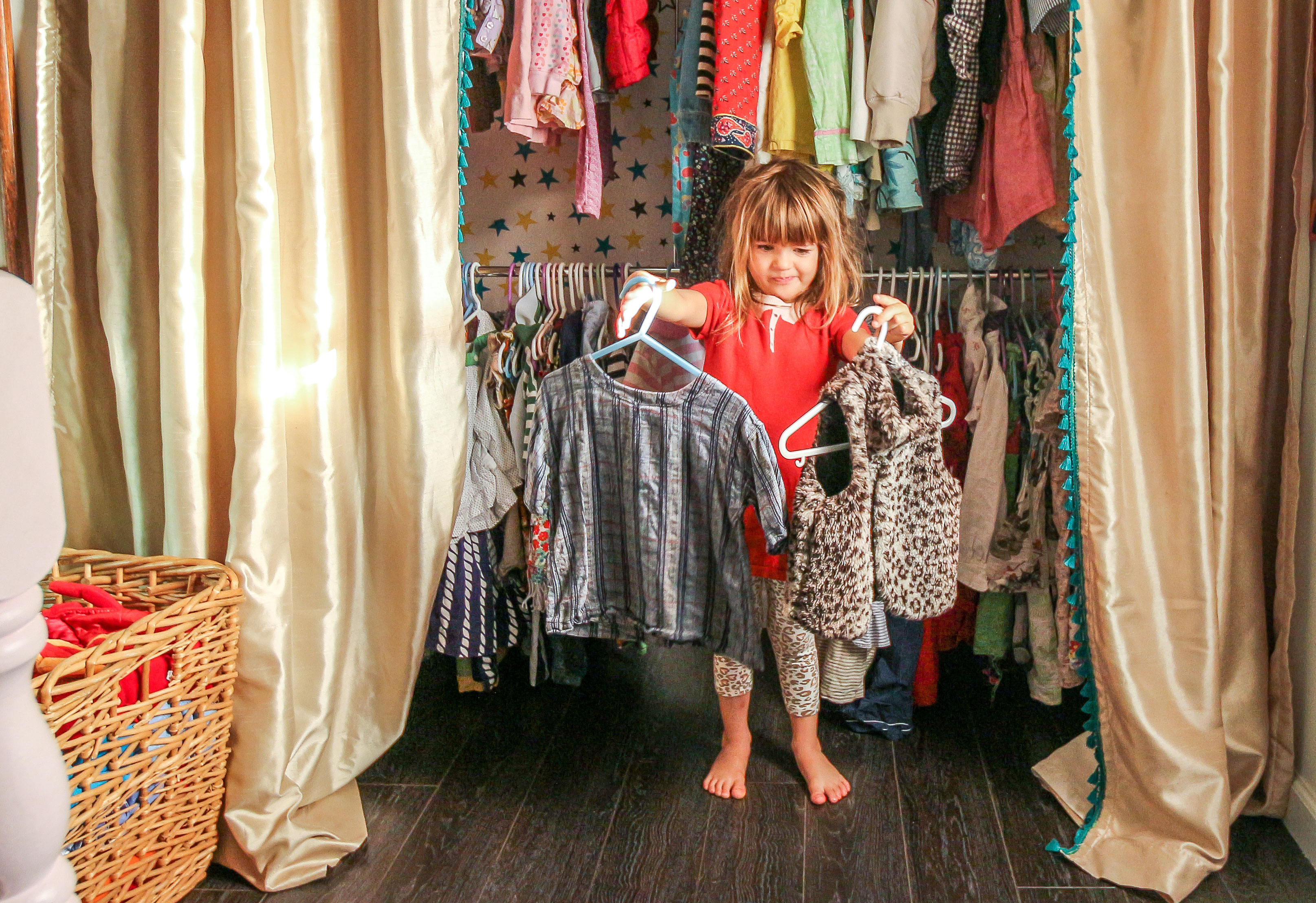 A young girl stands in front of an open closet filled with colorful children's clothes, holding up two outfits on hangers.