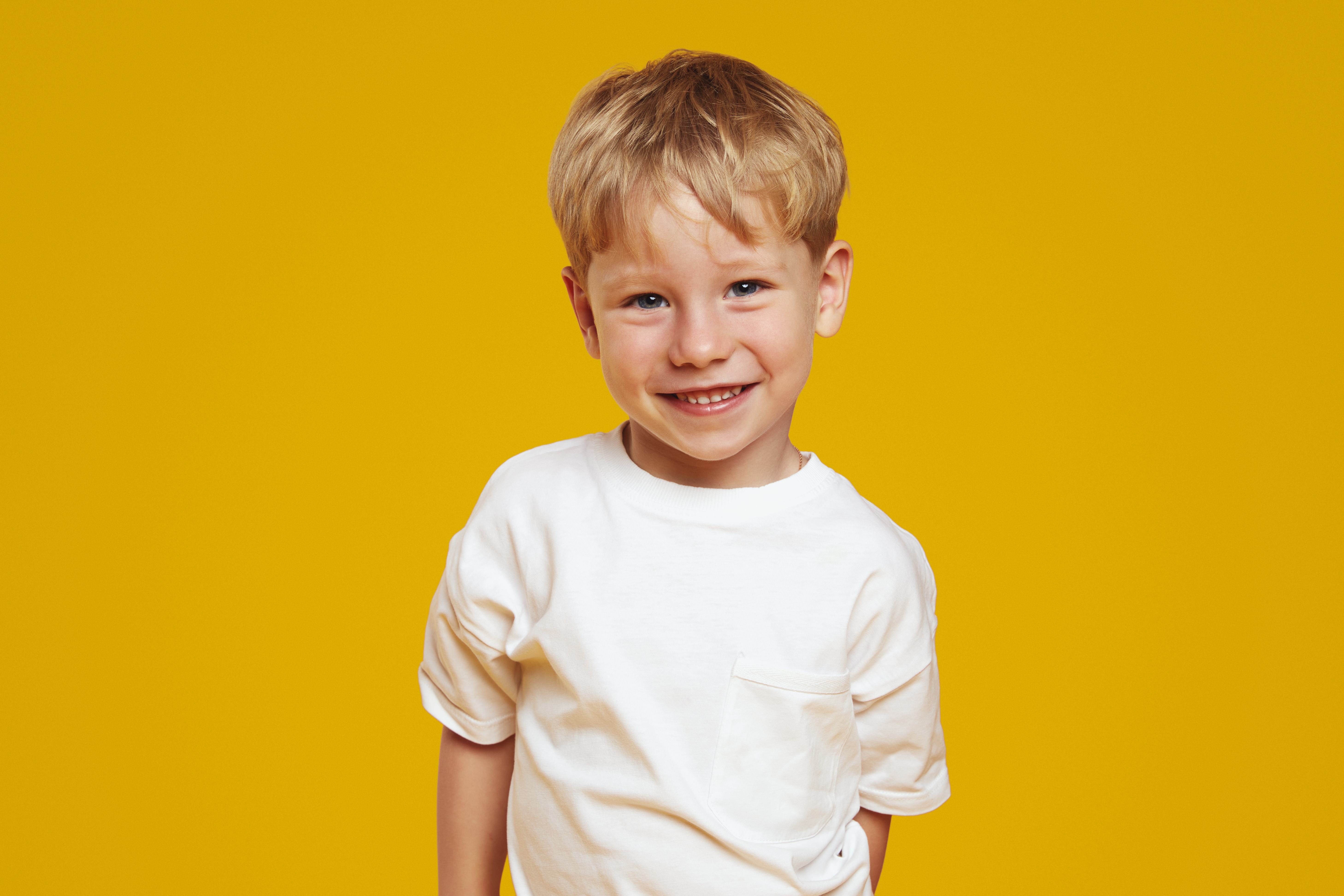 A young boy with blonde hair, wearing a white T-shirt, smiles against a bright yellow background.