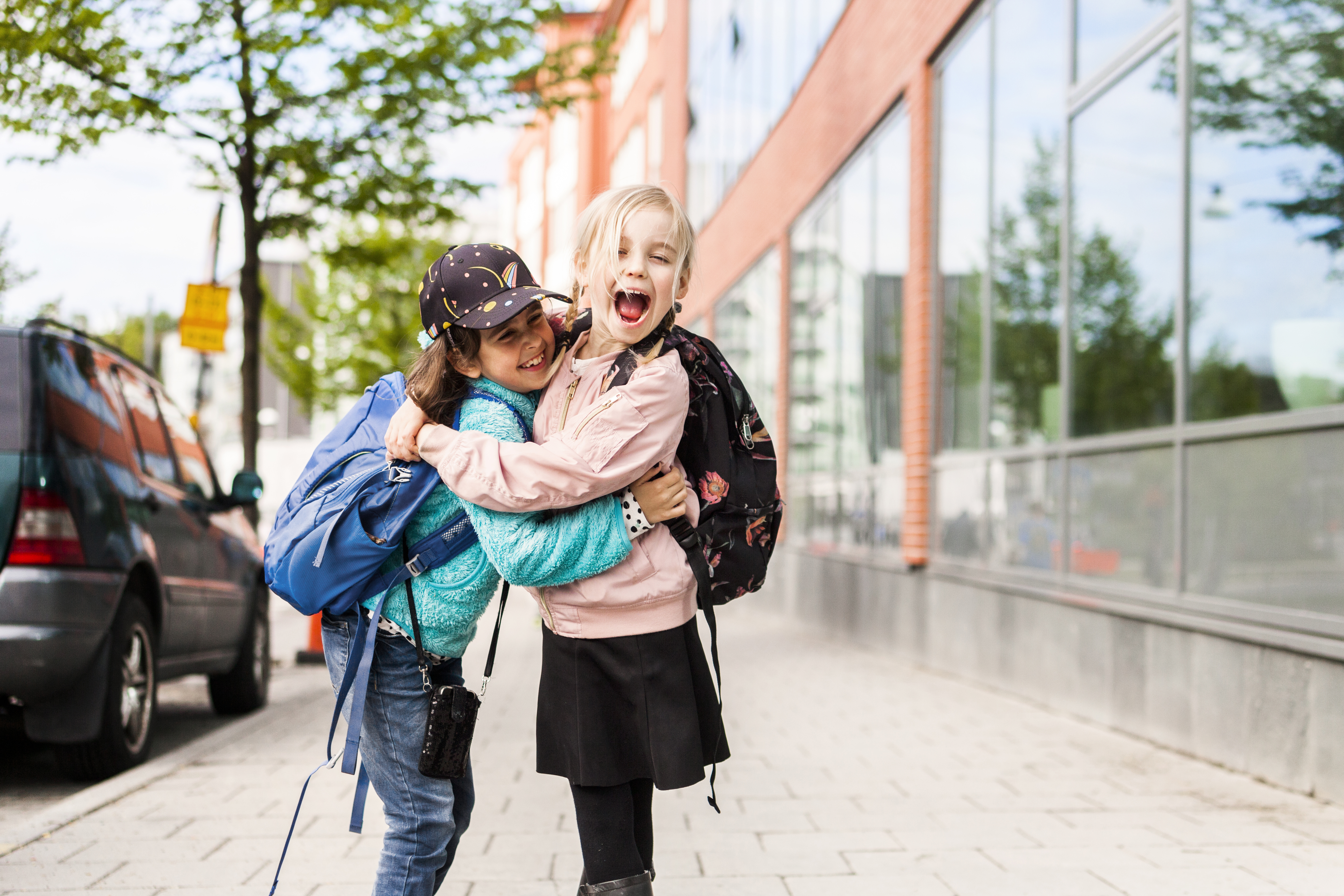 Two young children with backpacks hug joyfully on a city sidewalk, smiling and laughing.