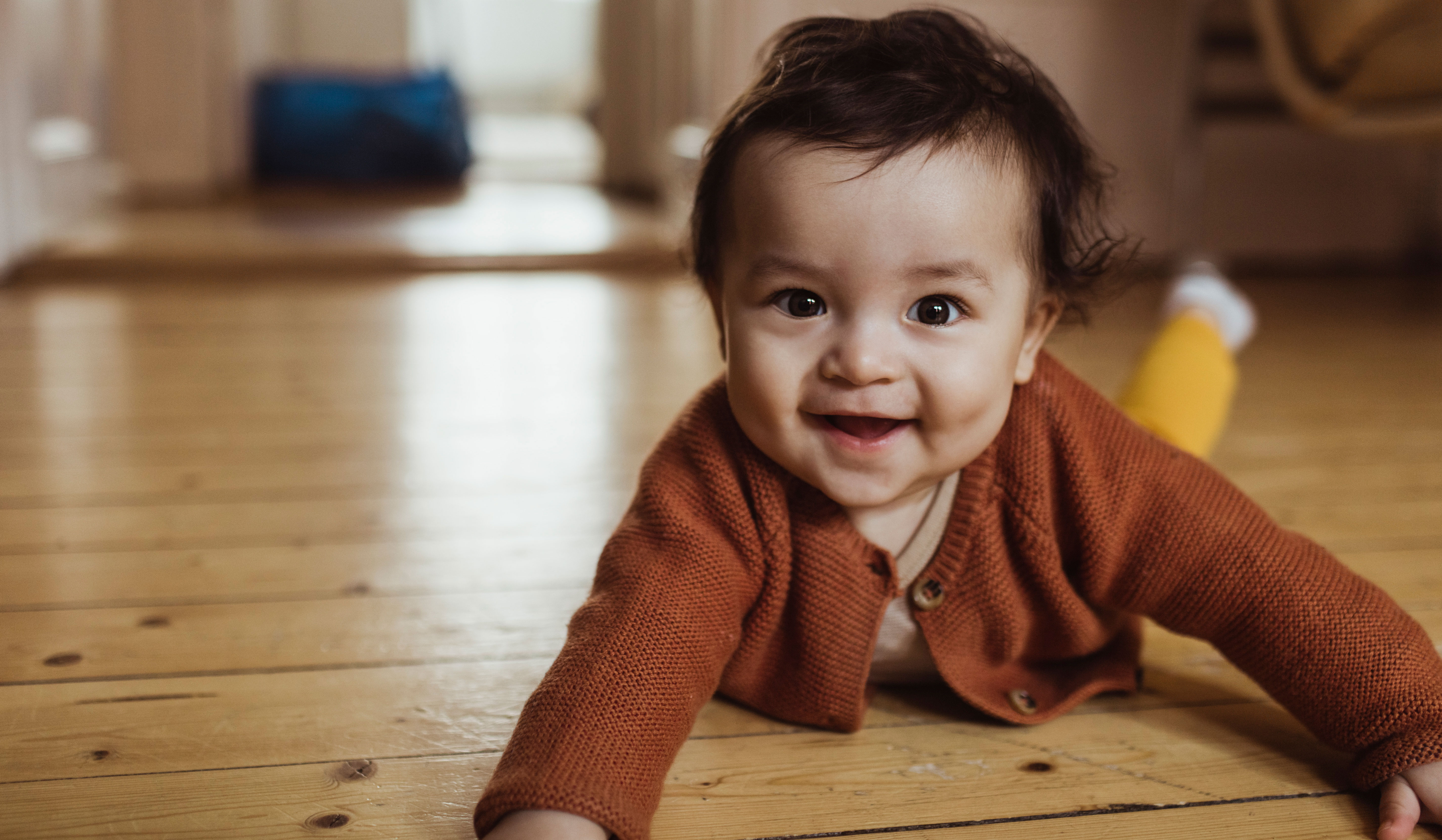 A smiling baby is lying on a wooden floor, wearing an orange knitted cardigan. The baby has dark hair and bright eyes, and appears happy and playful.