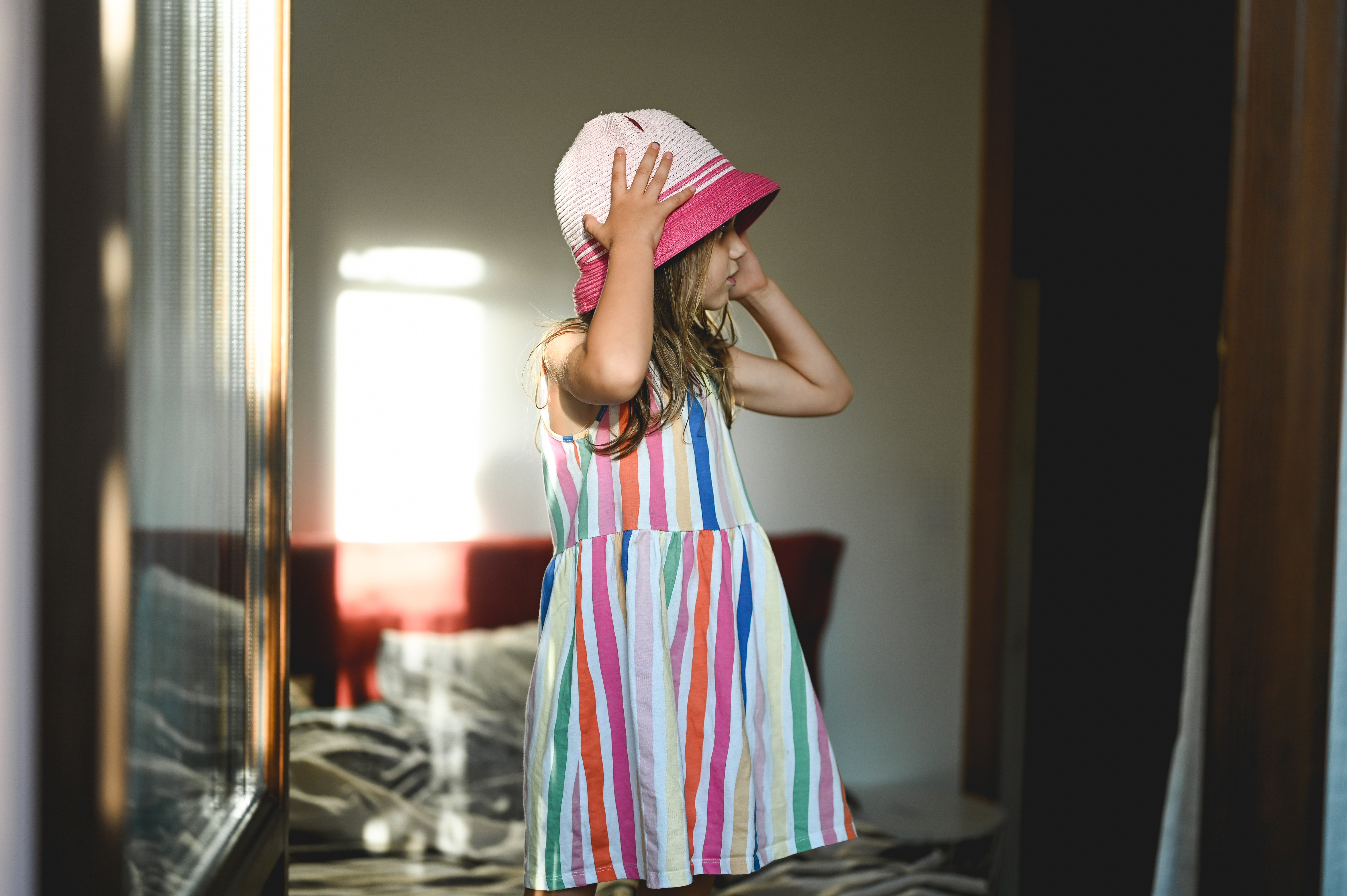 A young girl wearing a colorful striped dress and a pink hat stands in a room, adjusting her hat with both hands. She is facing sideways, with light streaming in from a window, casting a shadow on the wall behind her. The background includes a bed with a red headboard and some bedding.