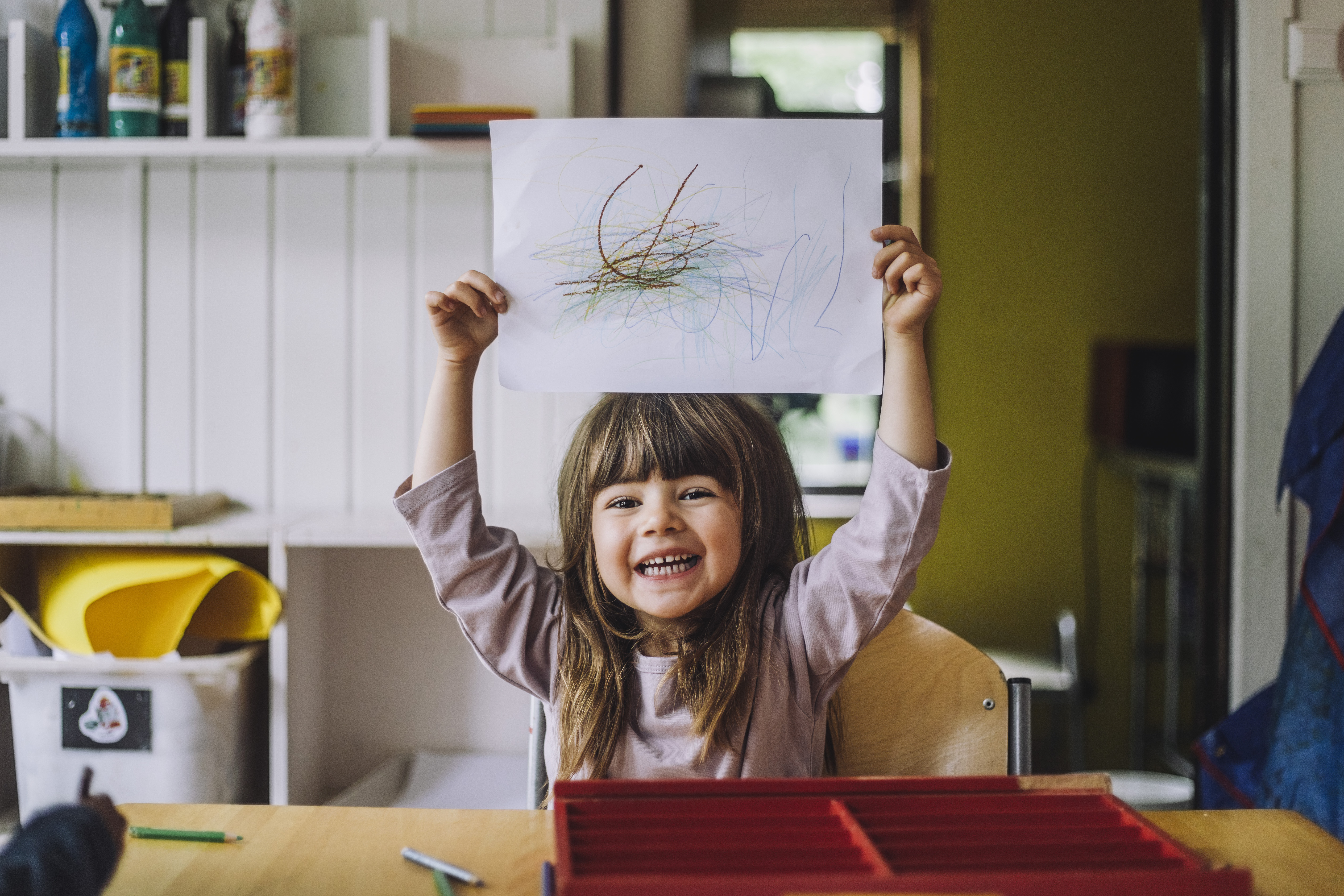 A smiling child with long brown hair holds up a paper with colorful scribbles, sitting at a table in a room with art supplies.
