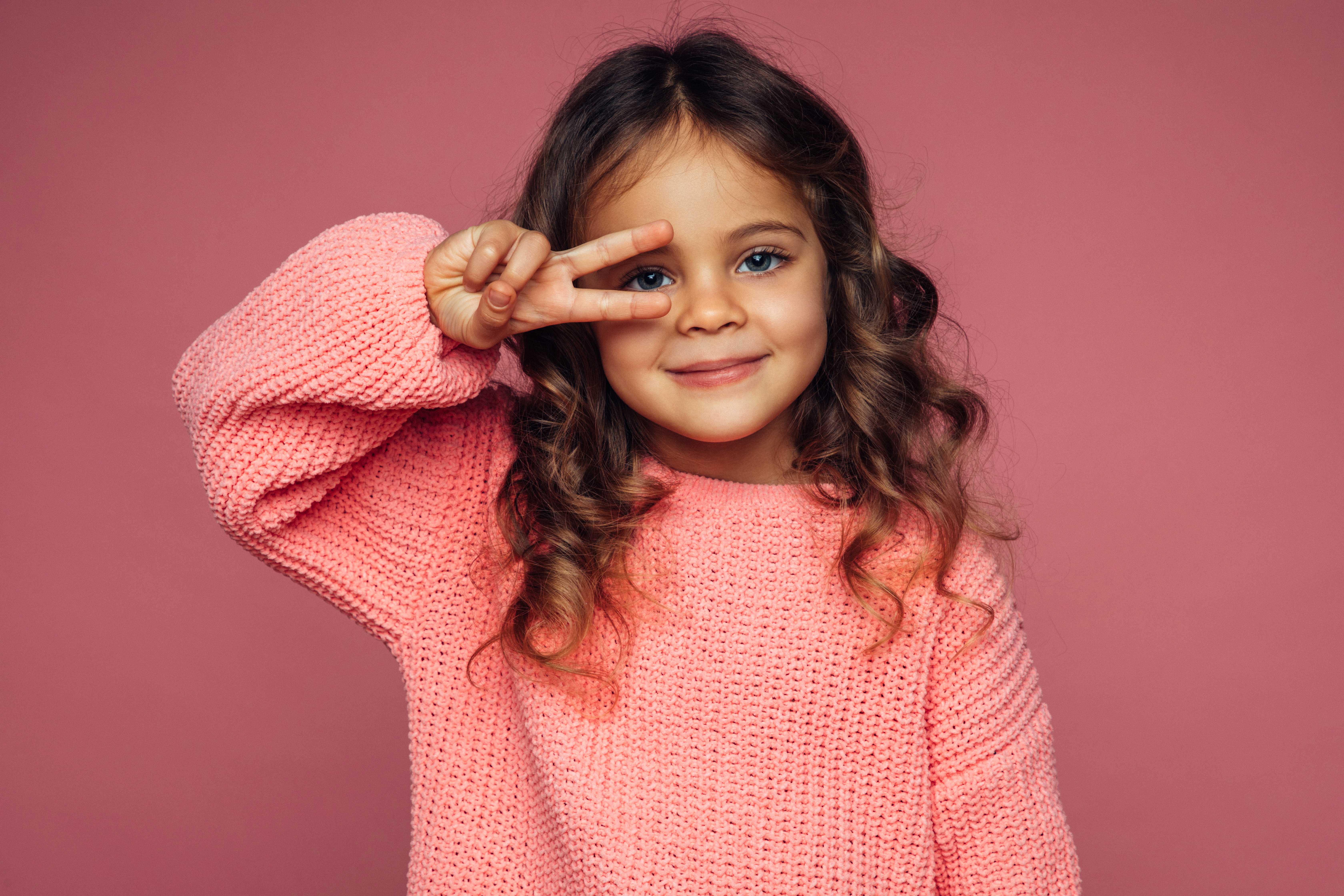 A young girl wearing a pink knitted sweater poses in front of a pink background, making a peace sign with her fingers near her eye and smiling.