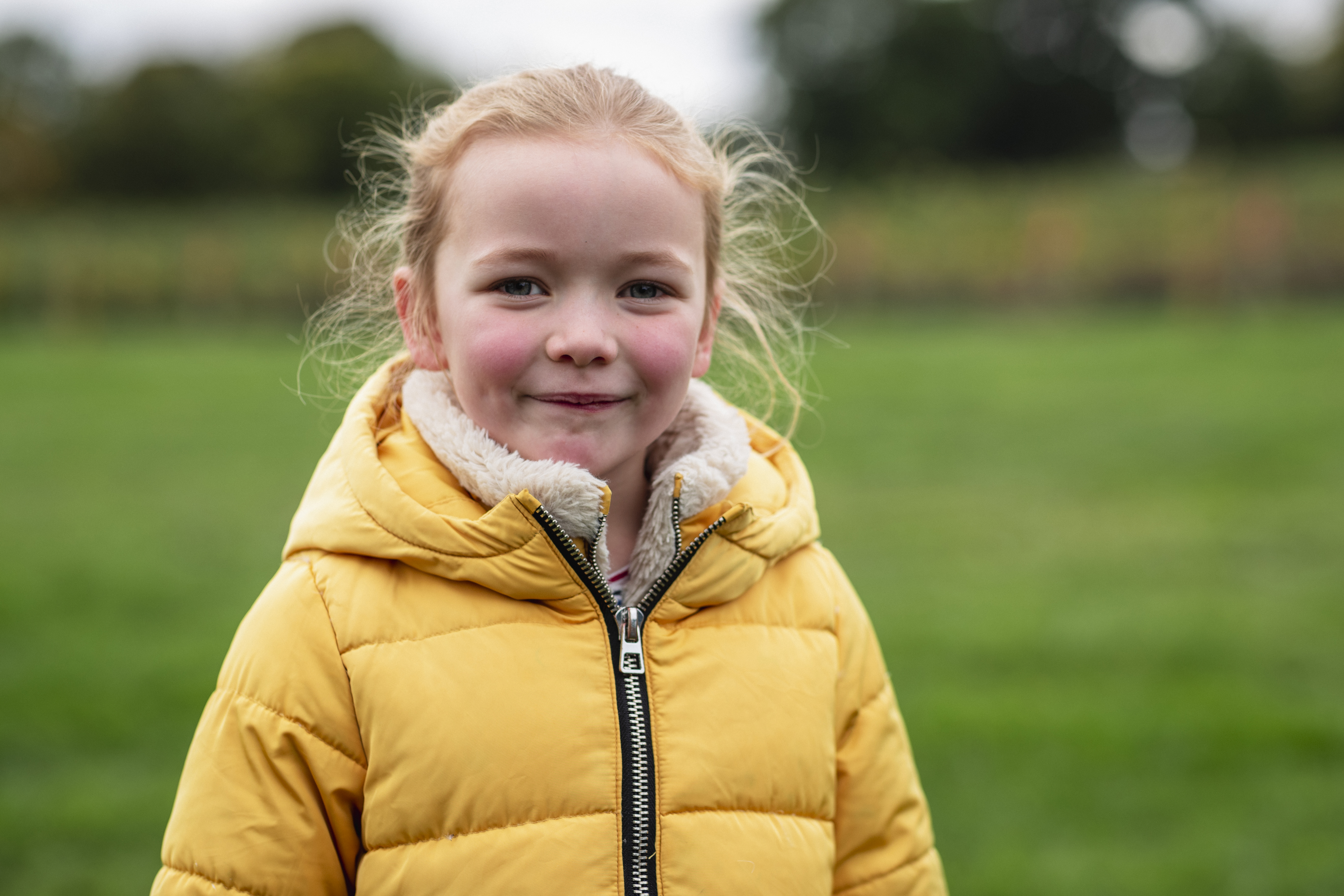 A young girl wearing a yellow puffy jacket stands outdoors, smiling with a grassy field and trees blurred in the background.