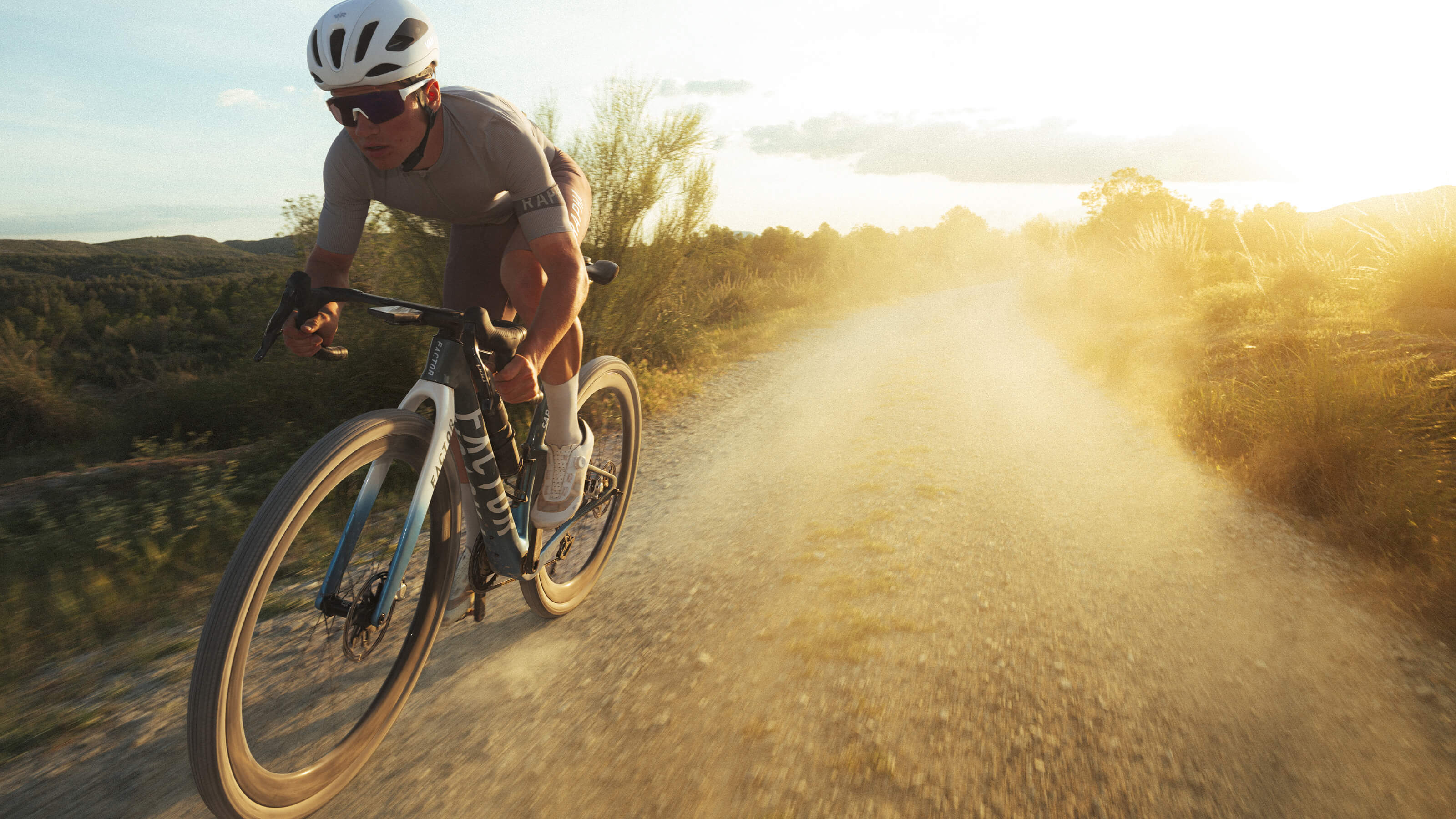 Active shot of a cyclist racing on the SARANA over a sun-drenched gravel road with a partly cloudy sky.