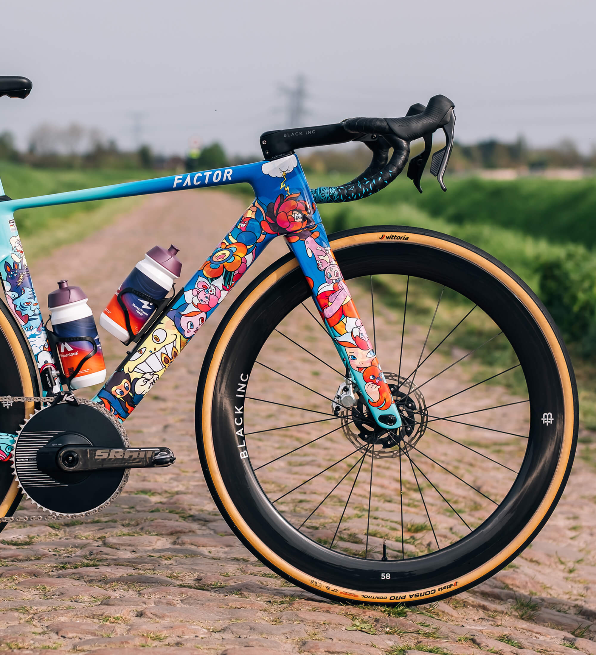 Medium shot of the front end of the drivetrain side of the MONZA VEXX Edition. The front wheel, handlebars, and front triangle are visible with a cobble road stretching deep into the background. The sun is shining, though the skies look gloomy.