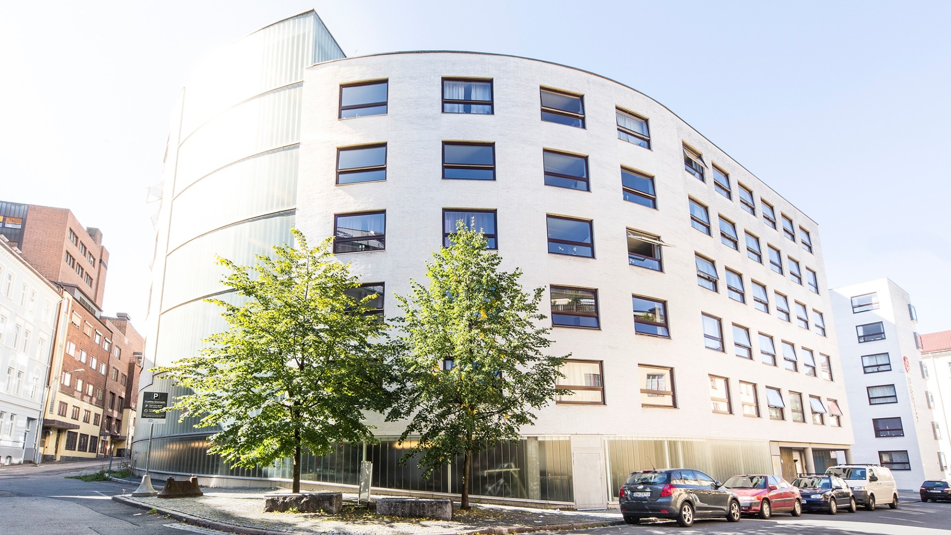 A picture of St. Hanshaugen Student Village. The picture shows a white building with a rounded shape that follows the curve of the road outside. Parked cars line the road.