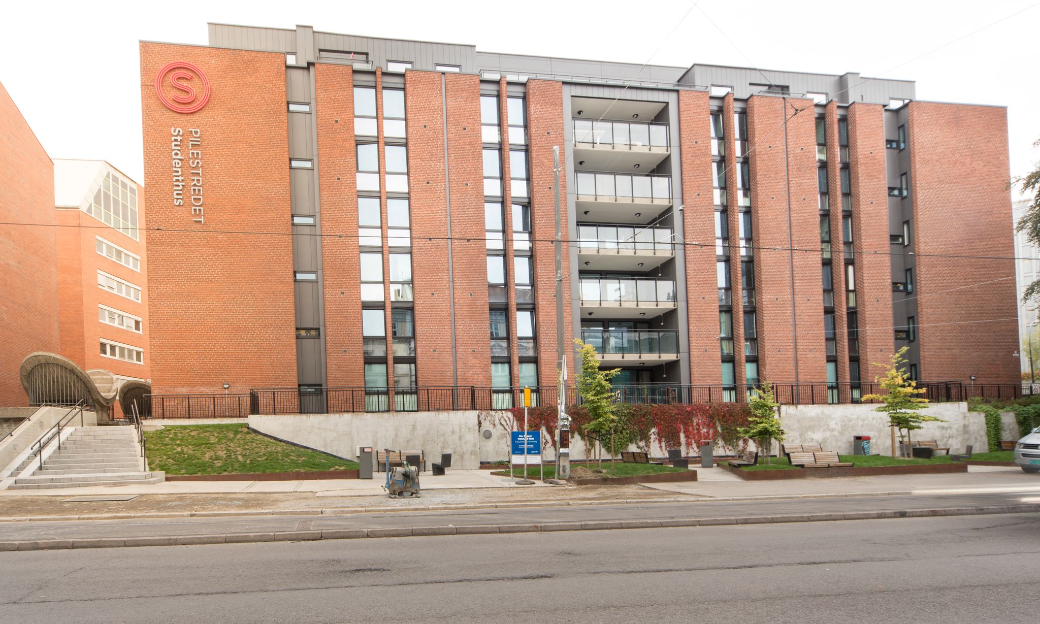 A picture of Pilestredet Student House. The picture shows a red brick high-rise building. In front of the Student House, there is a small area with seating and trees. There is a staircase leading up to the entrance of the Student House. The Student House is located by a road.