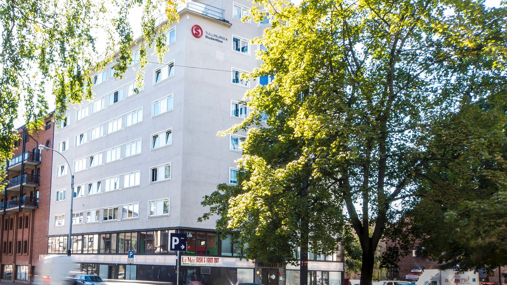 A picture of Tullinløkka Student Village. The picture shows a white building with a rooftop terrace. Alongside the building, there is a busy road.
