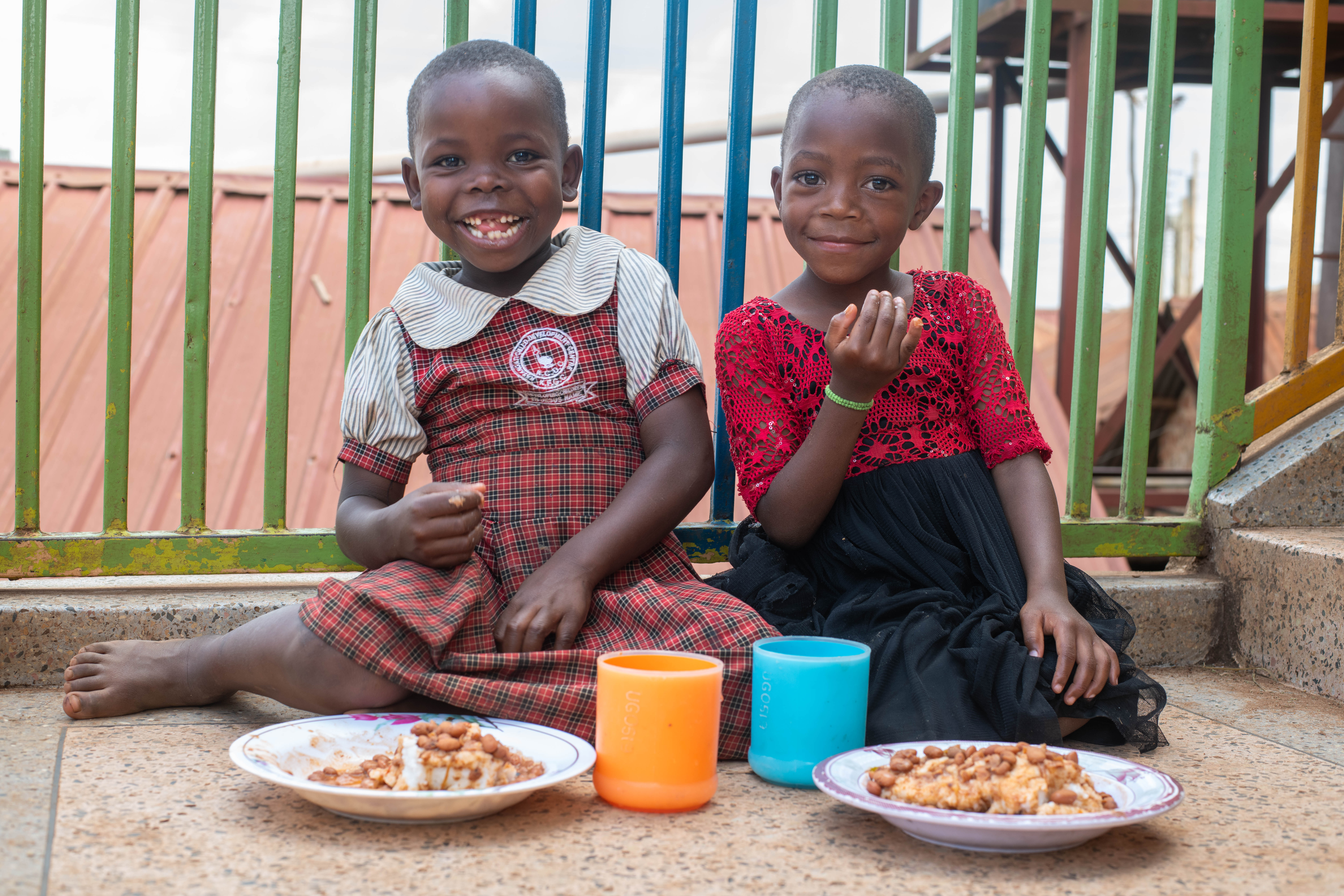 Two smiling children sit next to each other eating food from bowls.