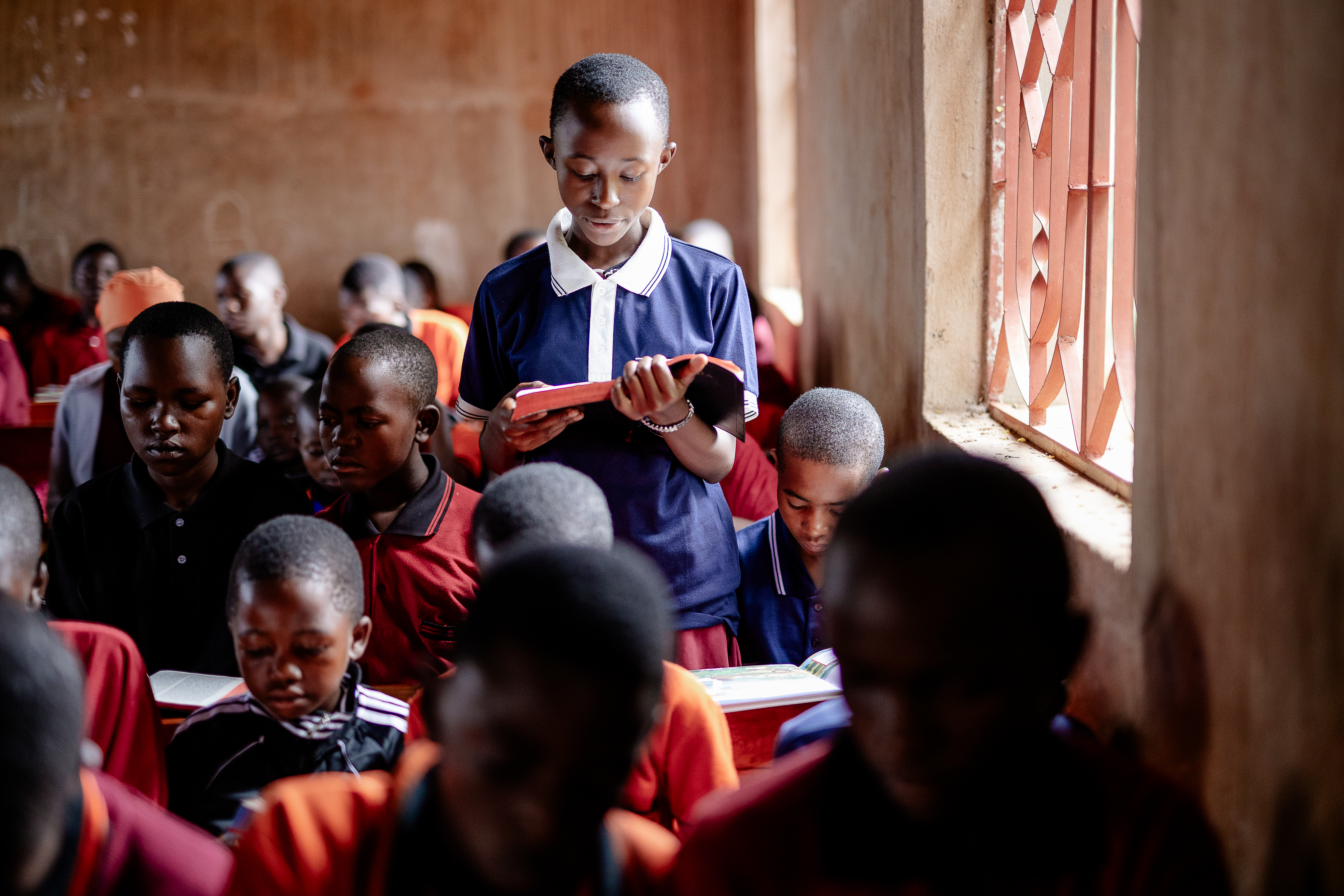 A youth in a blue and white top stands by a window, reading their Bible to a classroom full of children and youth.