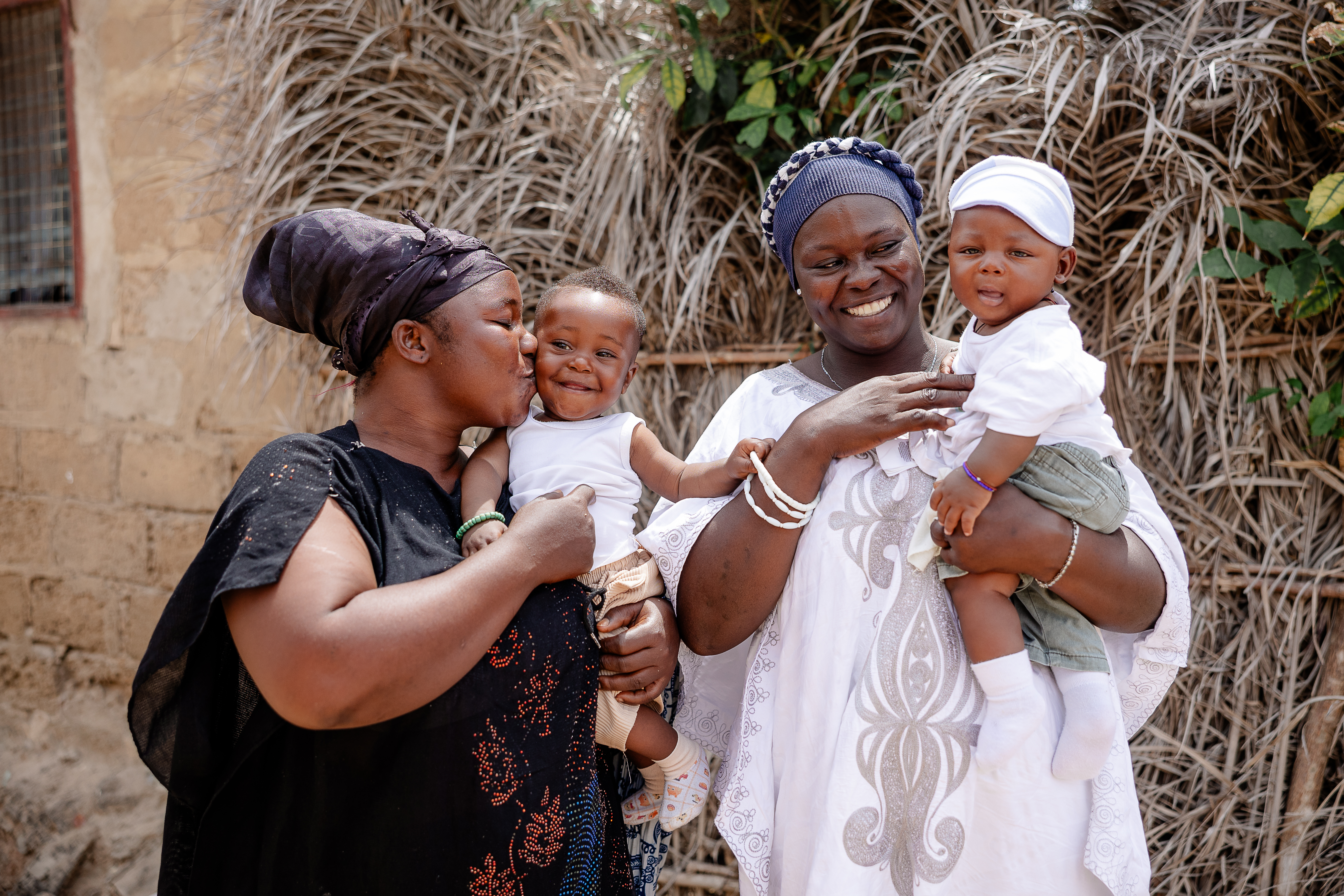 Two Ghanaian moms stand in front of a stone house each holding their baby boys in their arms.