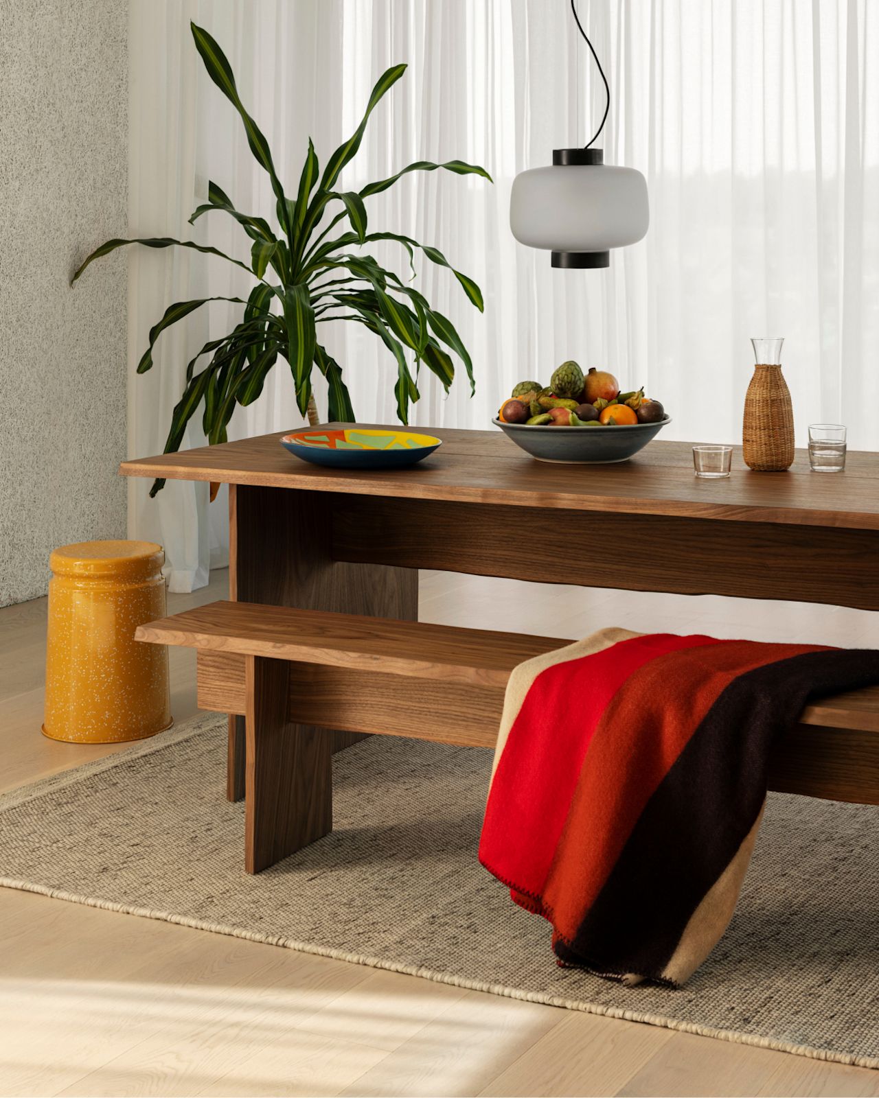 Hem - A dining room scene featuring a Bookmatch Table + Benches Set in Walnut with a Dusk Lamp above and Rope Rug Natural underneath. A Block Throw in the shade Fire is draped over one of the benches and a Last Stool Splatter (Yellow / Off-White) stands beside.