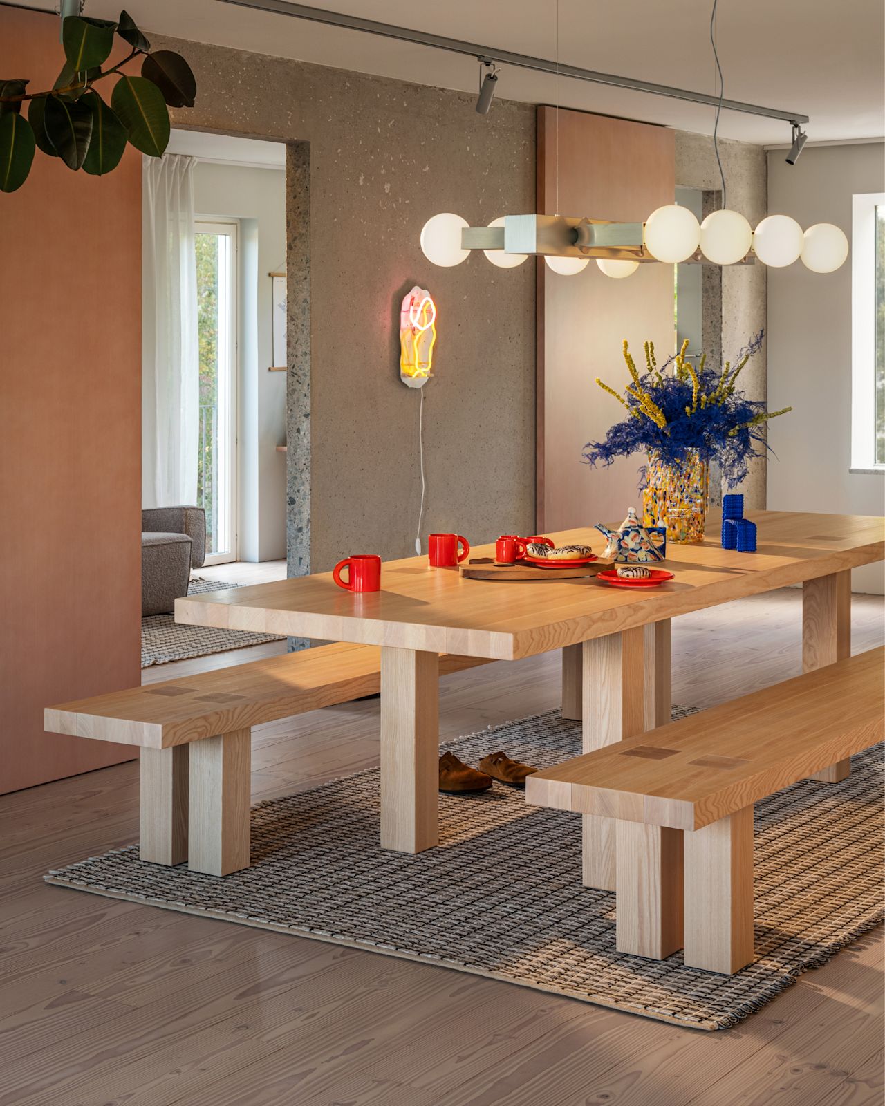 A lifestyle image of a dining scene featuring Max Table + Benches Set, Rope Rug, Knuckle Linear Chandelier, Globos Neon, Bronto Tableware, and Brute Vase.