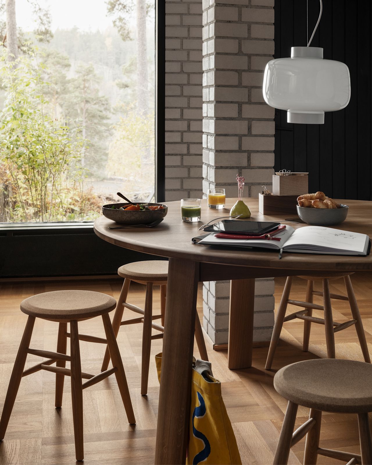 A lifestyle image of a dining scene featuring Alle Round Table, Drifted Stools, and Dusk Lamp.