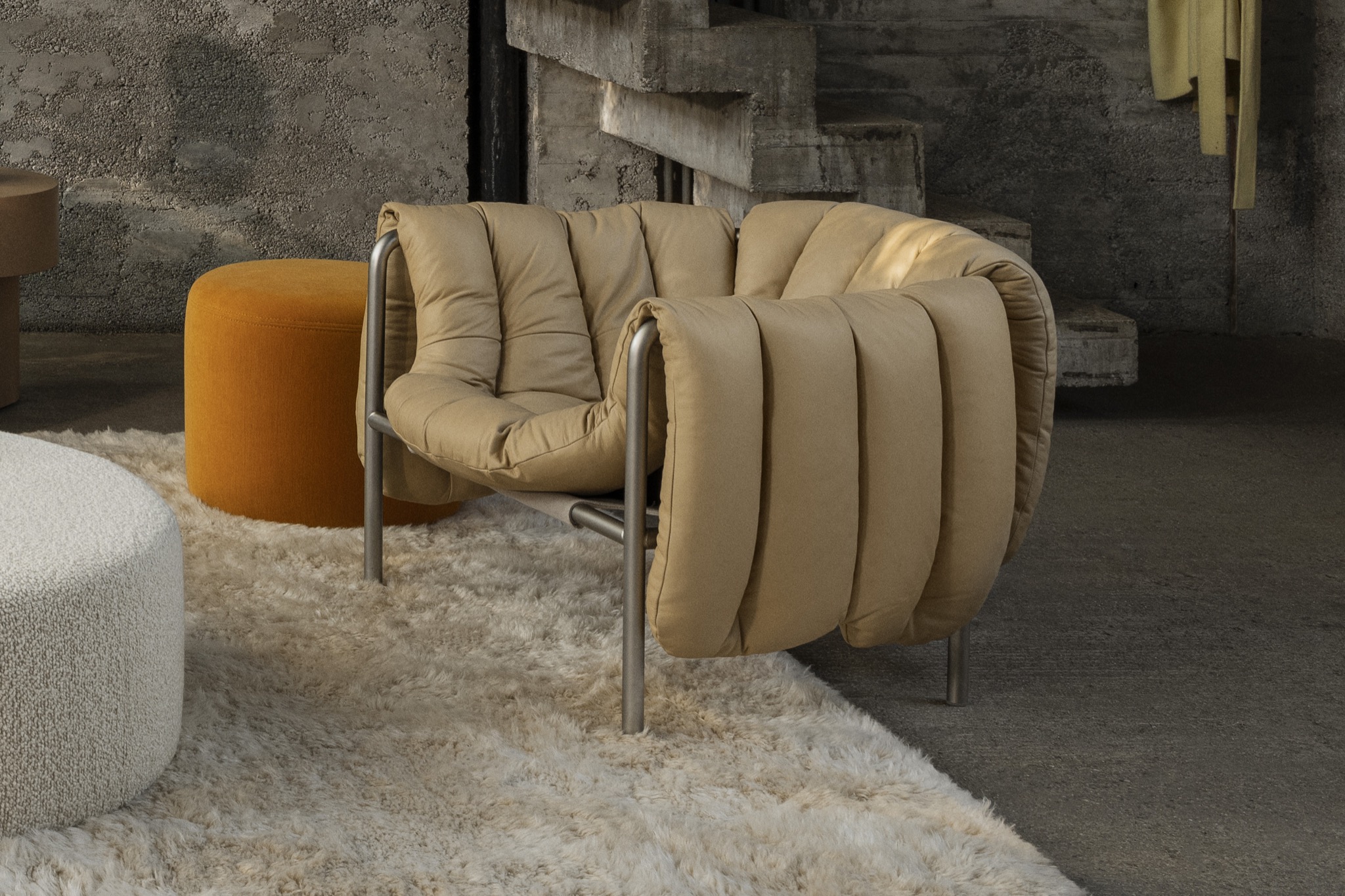 Hem - A living room scene featuring a Puffy Lounge Chair in Sand Soft Leather / Stainless Steel on top of a Monster Rug in Beige / Off-White. In the background is a Bon Pouf Round in Ochre and a Bon Pouf Round Large in Eggshell.