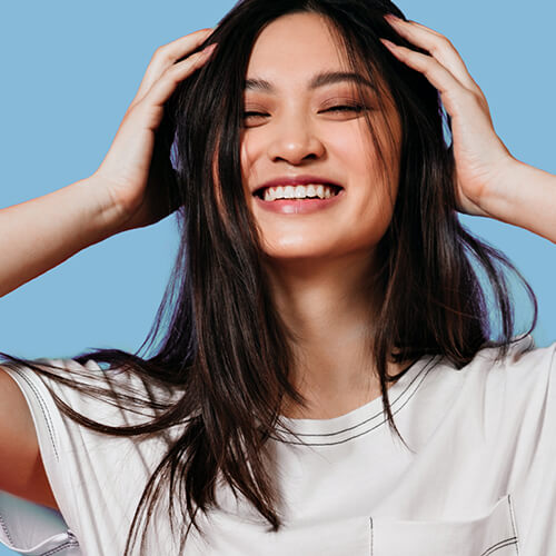 A young woman of mixed descent smiles while using dry shampoo on her long, fine, brown hair.