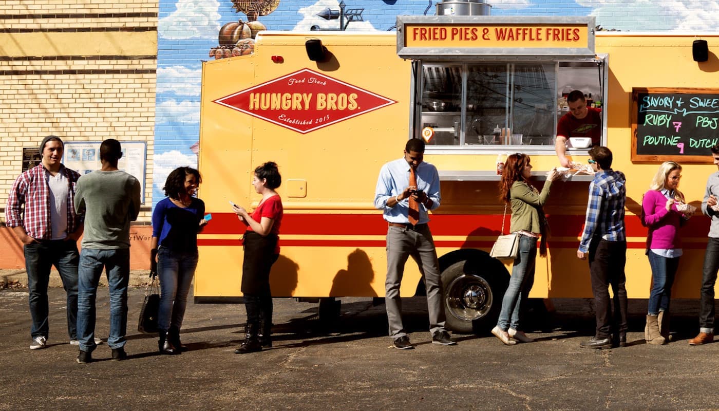 Yellow and red “Hungry Bros.” truck by brick building; truck says “Fried Pies & Waffle Fries”; customers gather and order.