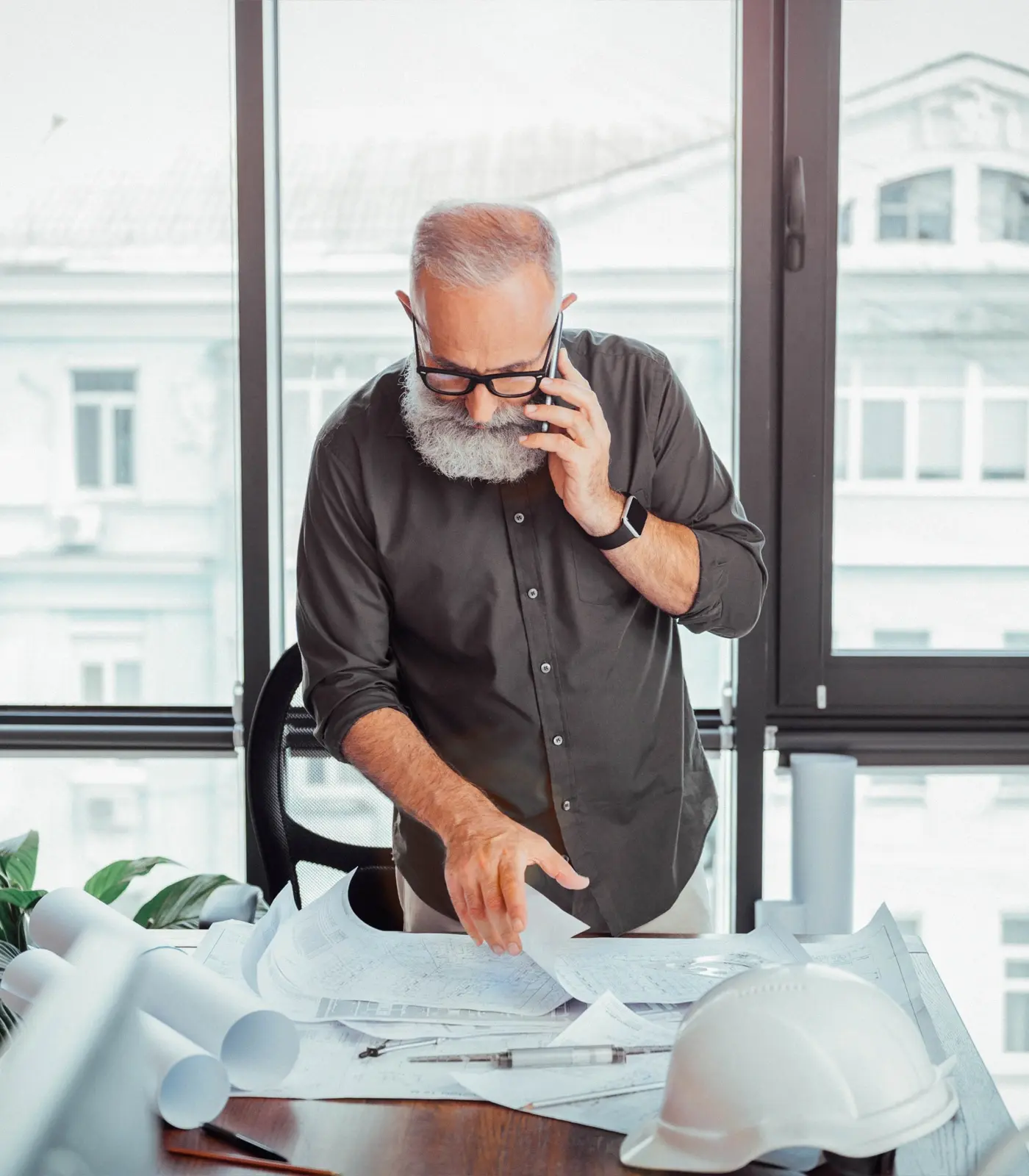 An architect working through building plans that are on a table in front of him while he is talking on the phone.