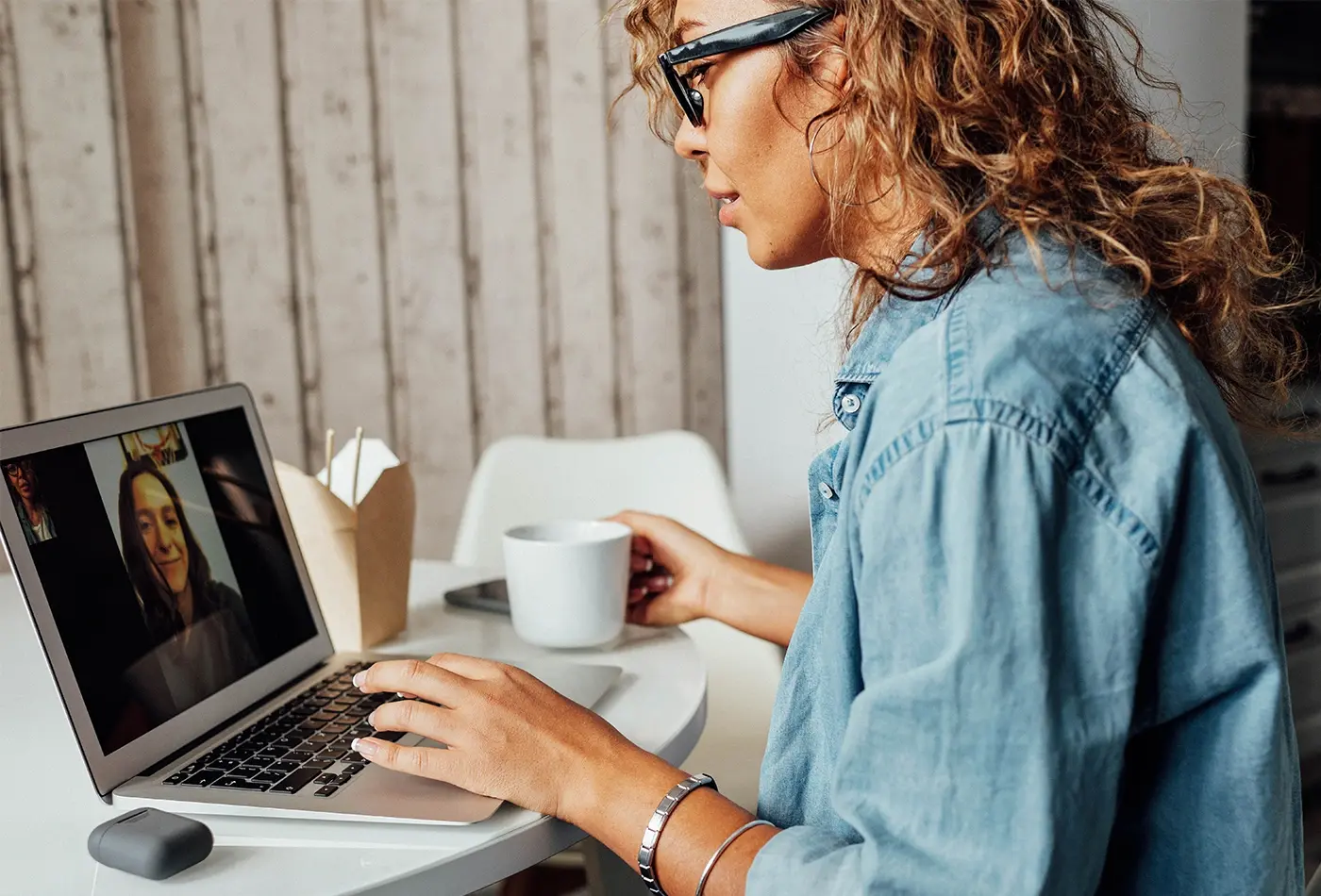 Woman at a table on a laptop video chatting with a woman. Her left hand is on the laptop while her other hand holds coffee.