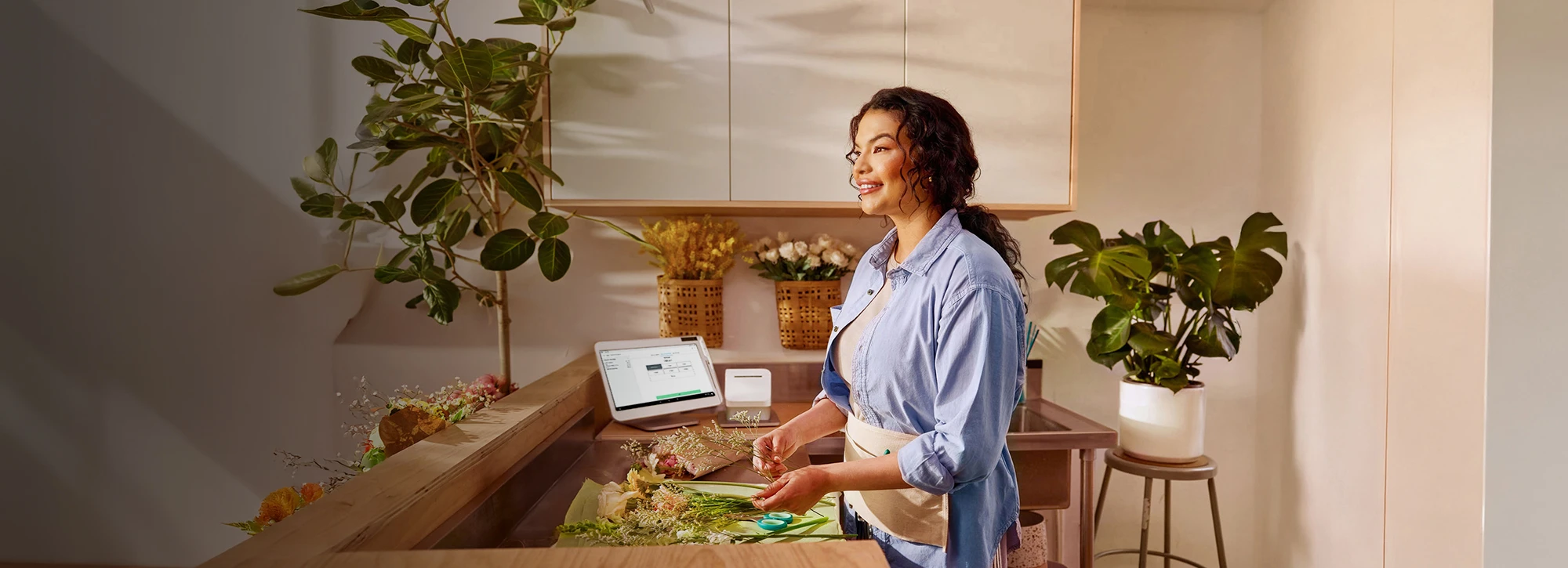 Smiling florist in blue shirt arranges bouquet behind counter; Clover Station Solo with payment screen sits nearby.
