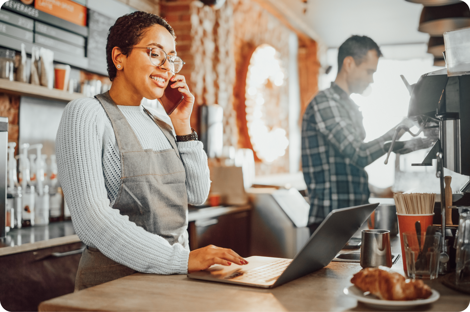 Coffee shop worker standing behind counter talking on the phone while working on a laptop.