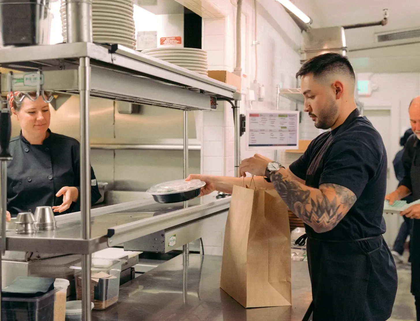 Chef standing in a kitchen handing a to-go order to a kitchen staff member who is packing the order into a paper to-go bag.