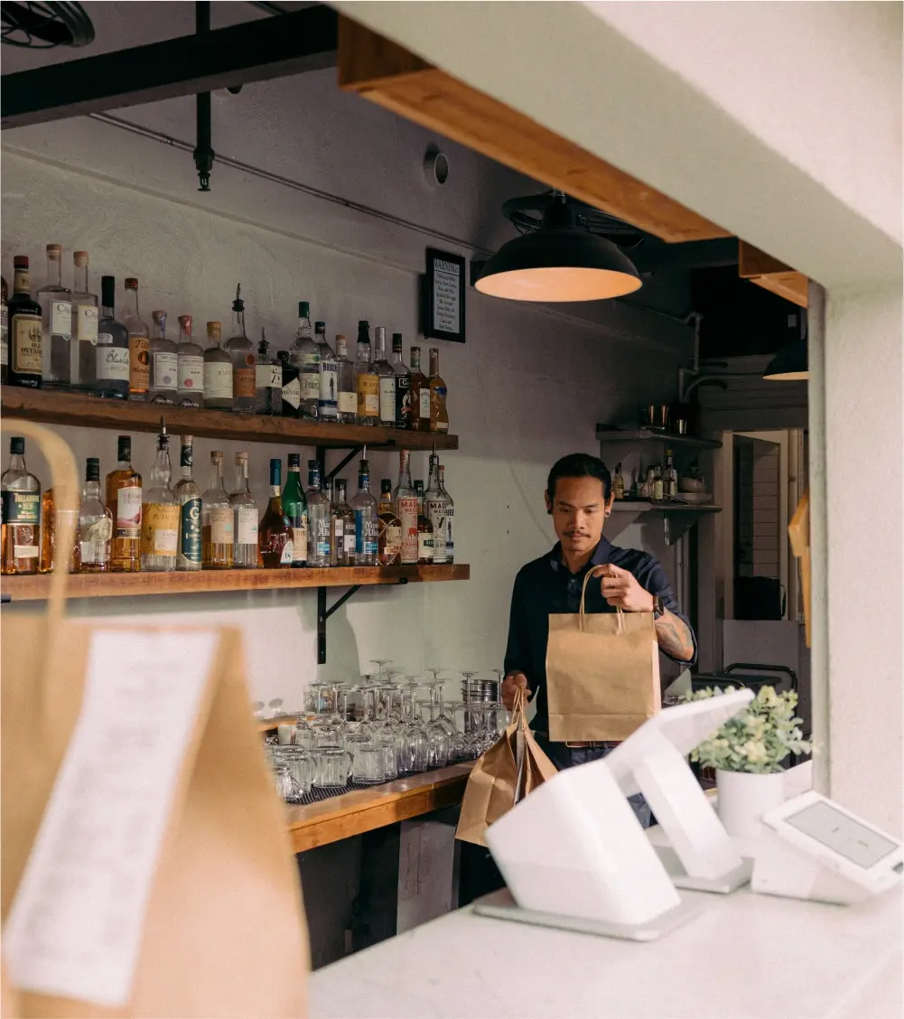 Restaurant worker behind a bar carrying prepared takeout orders to the register for pickup.