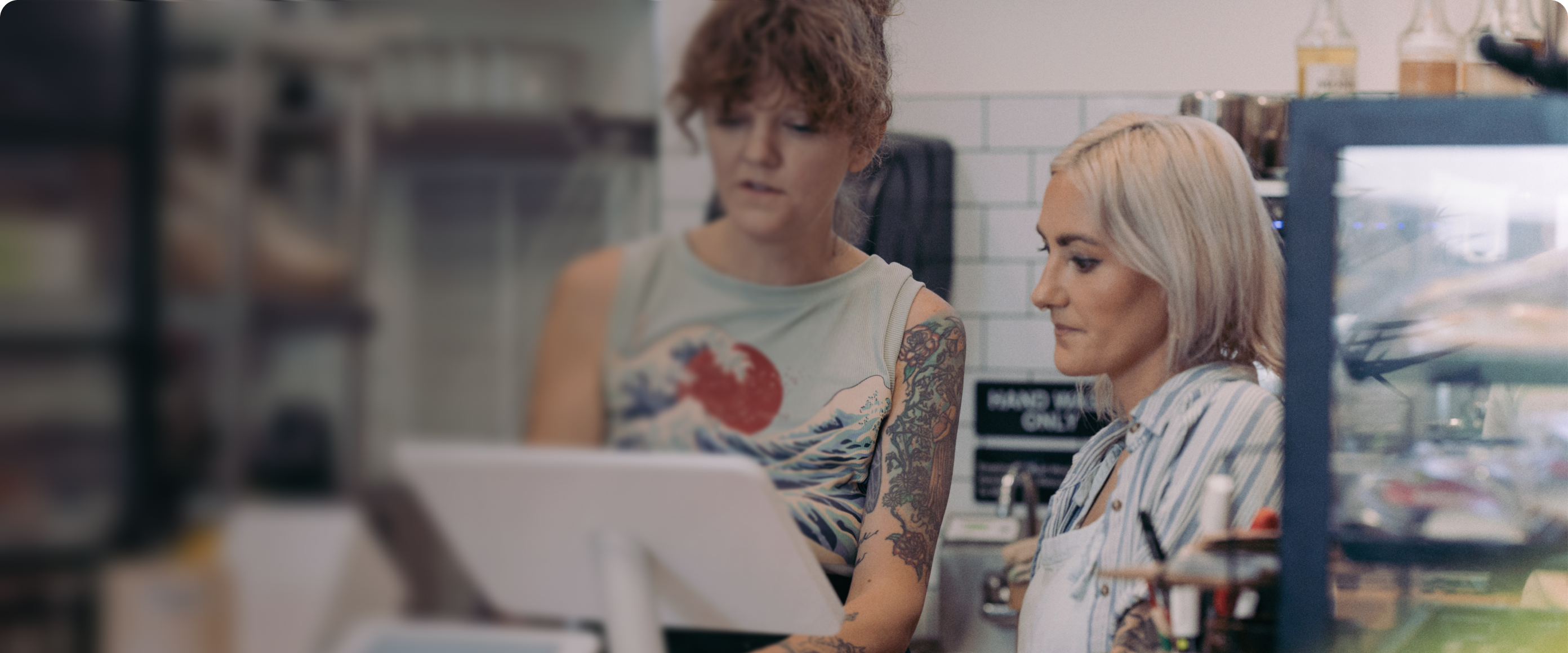 Two women at the counter viewing a 'Station Duo' POS system with a front facing 'Mini' device.