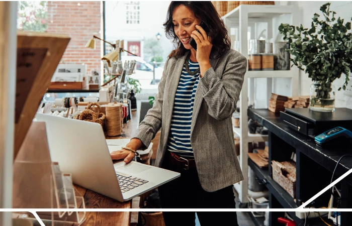 Sales woman dressed in business attire standing behind a counter talking on phone while using a laptop.