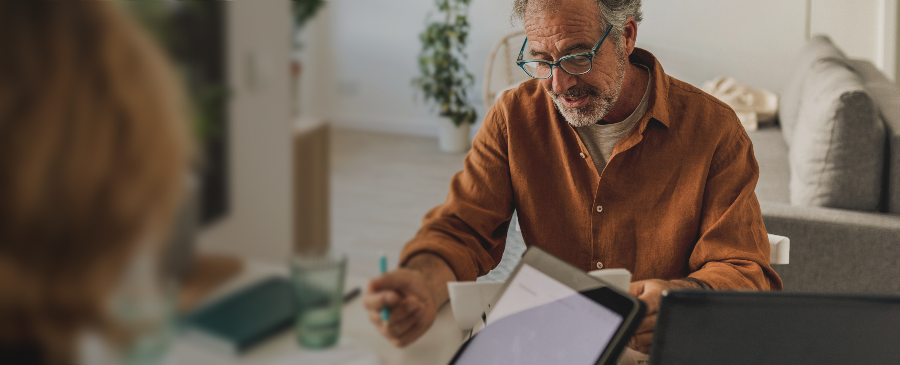 A man wearing brown shirt with some papers and a pen sitting at the desk explaining something to a person who is sitting opposite to him.