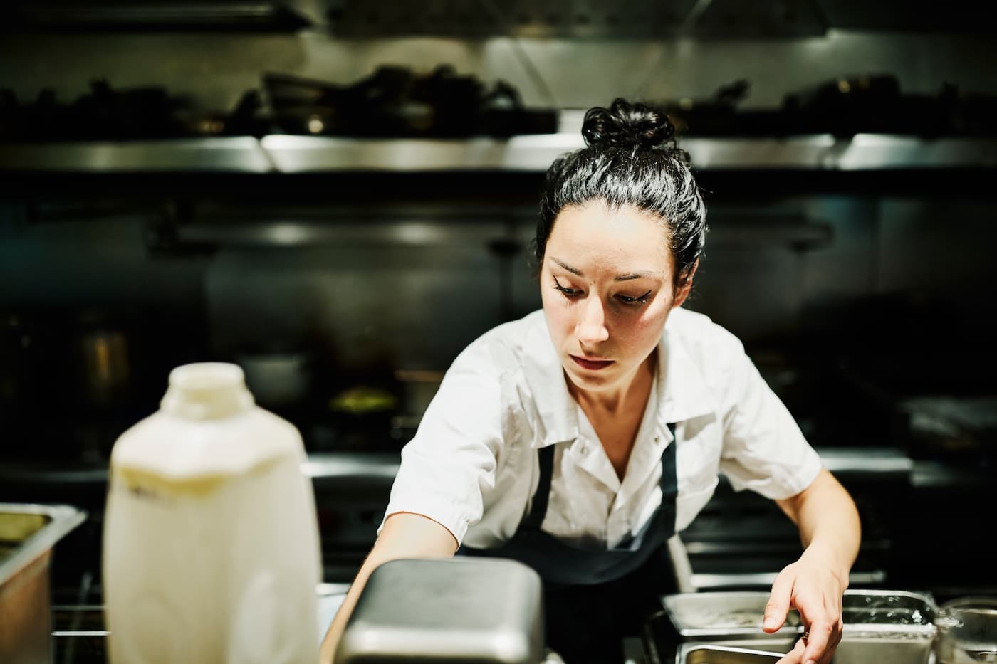 Chef in white shirt and apron works in commercial kitchen; food prep containers and stove visible in clean, focused setting.