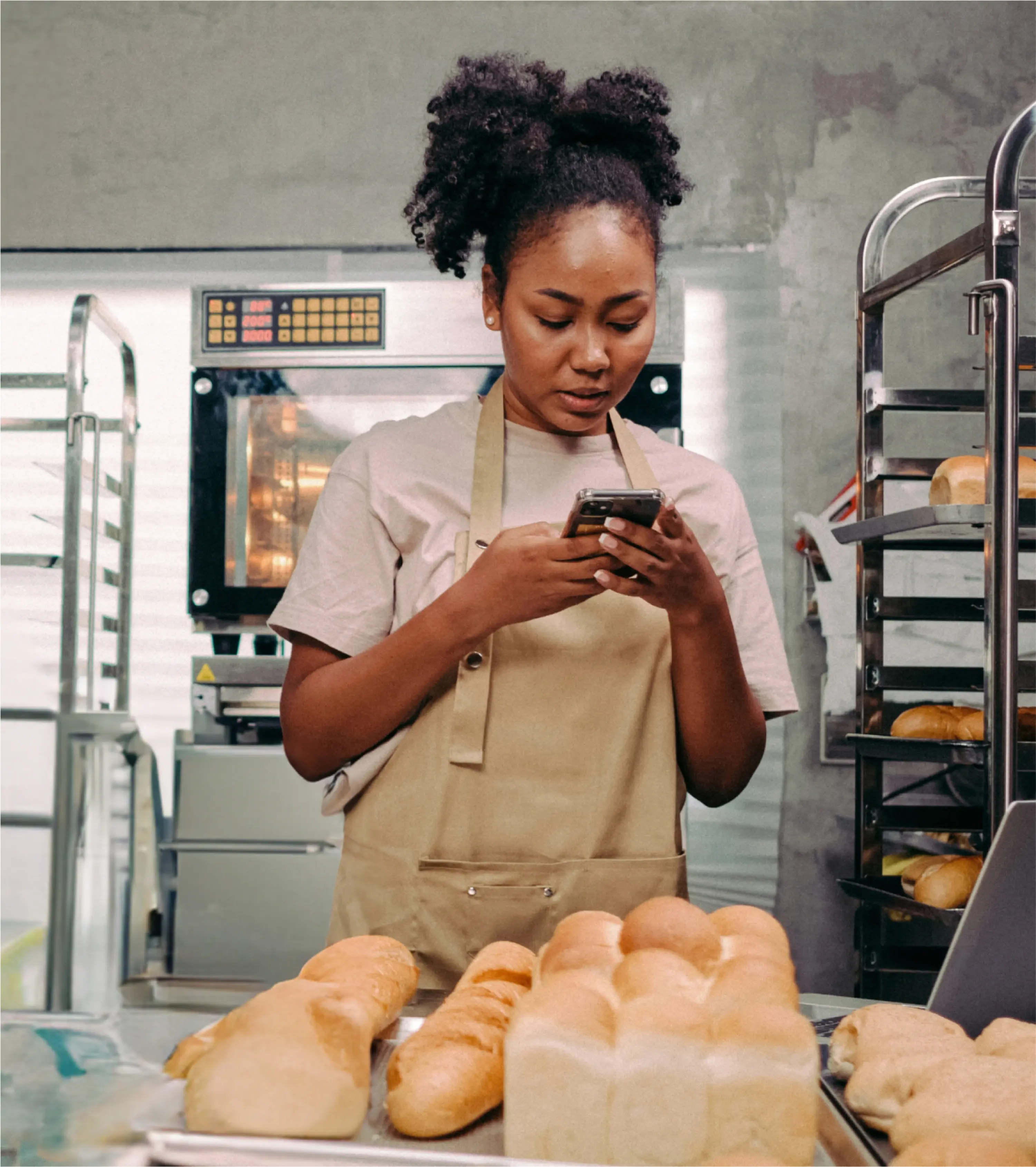 Woman standing in the kitchen of a bakery using her phone to take a picture of bread on a tray